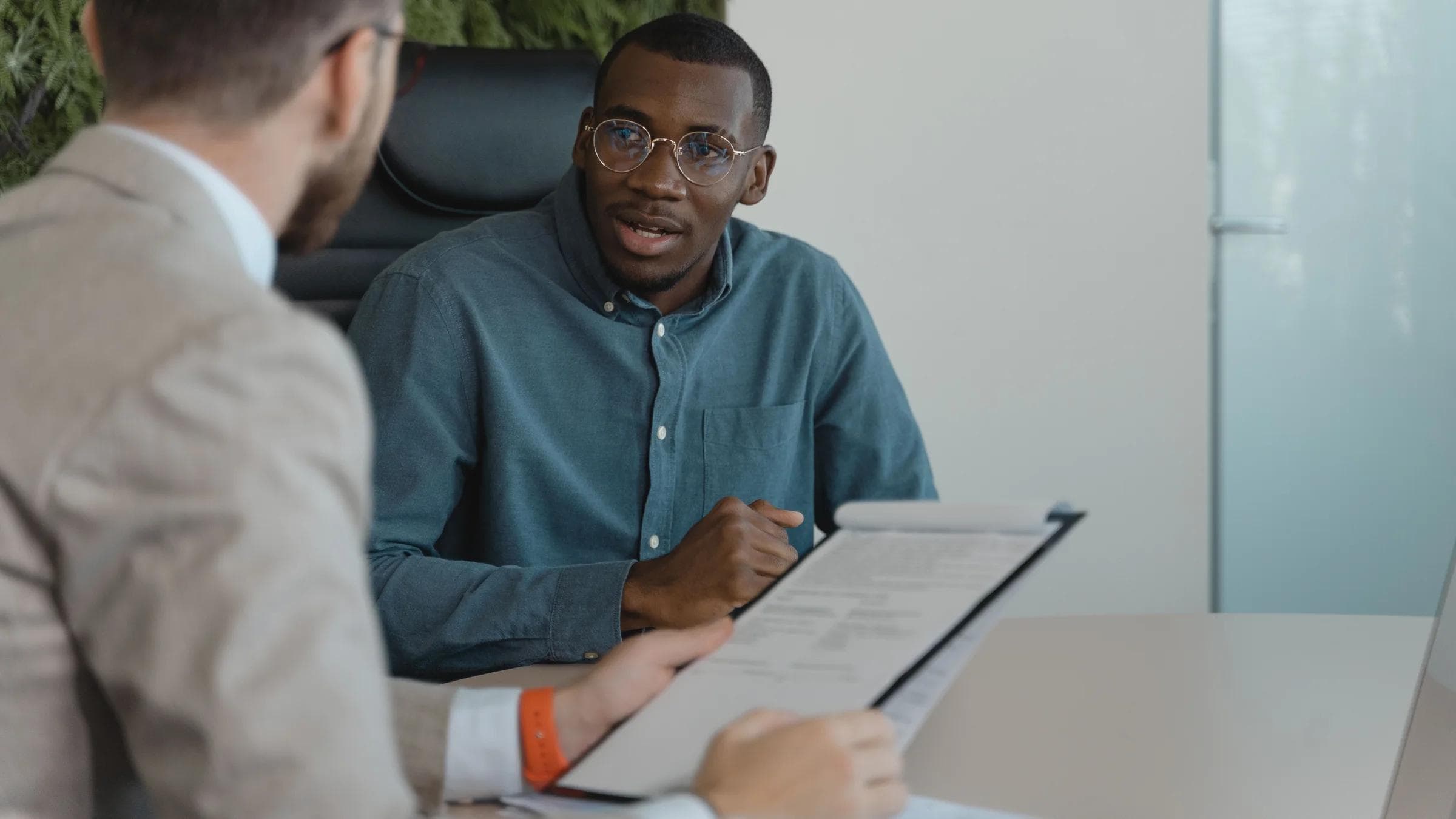 Two men engaged in a professional meeting at an office table with documents and a plant wall background.
