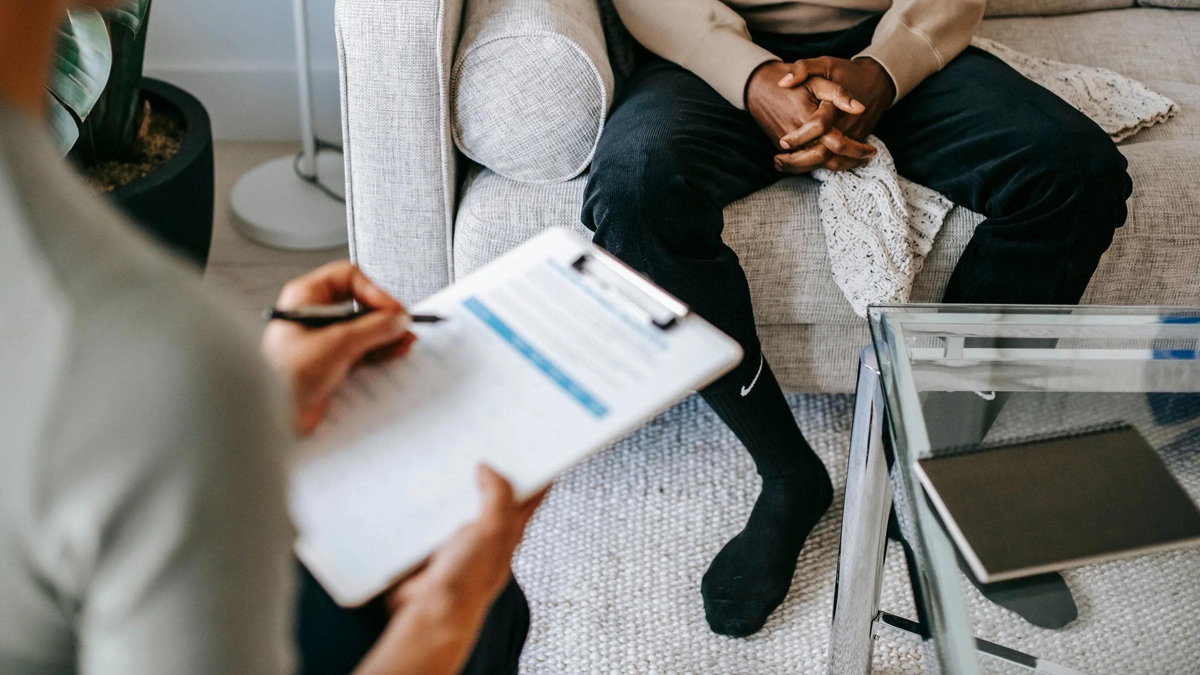 Blurred unrecognizable female therapist taking notes on clipboard while sitting in front of ethnic male person in clasped hands during meeting.