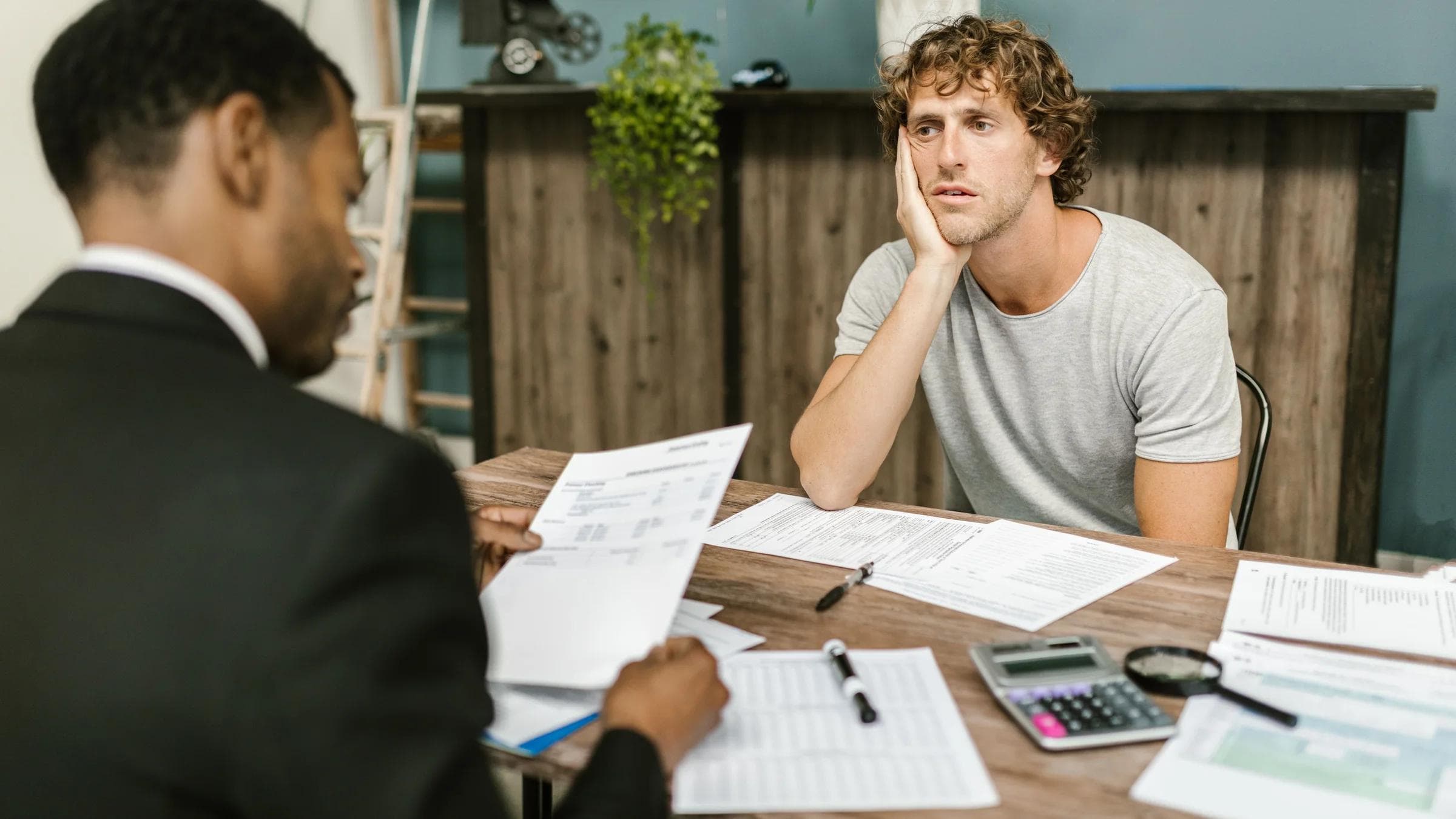 Man in office, appearing stressed during paperwork discussion, sitting opposite professional.