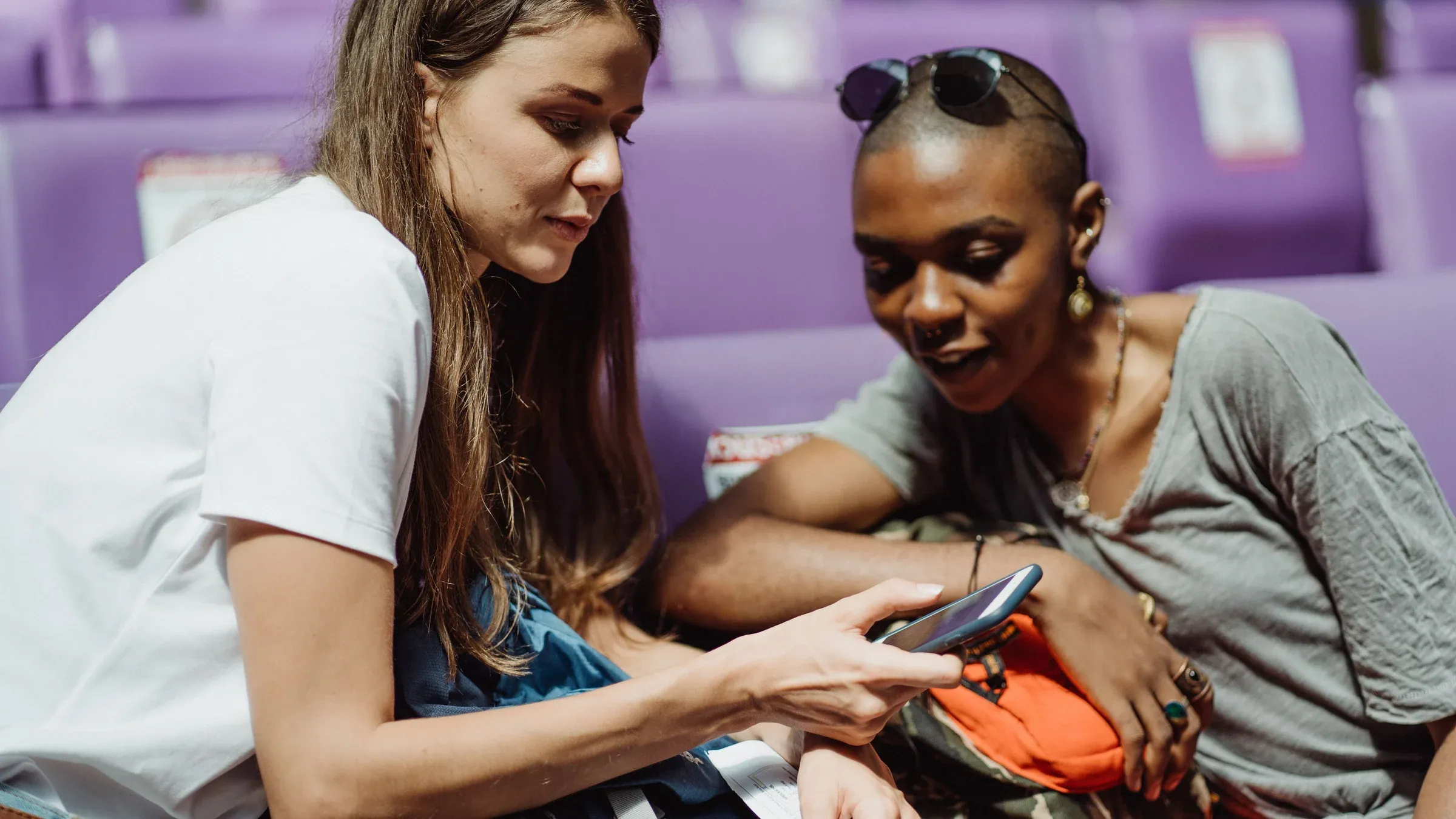 Two adult women sitting together and sharing a smartphone in a public waiting area with purple seats.