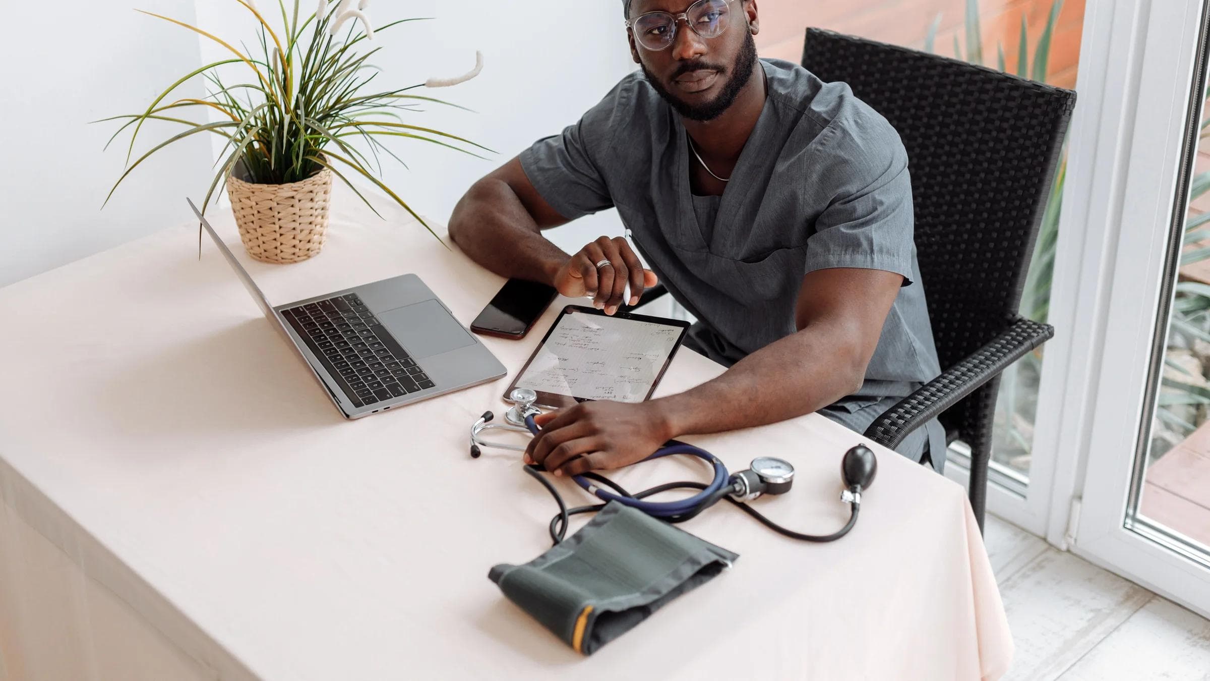 A doctor reviews records on a tablet beside a laptop in a medical office.