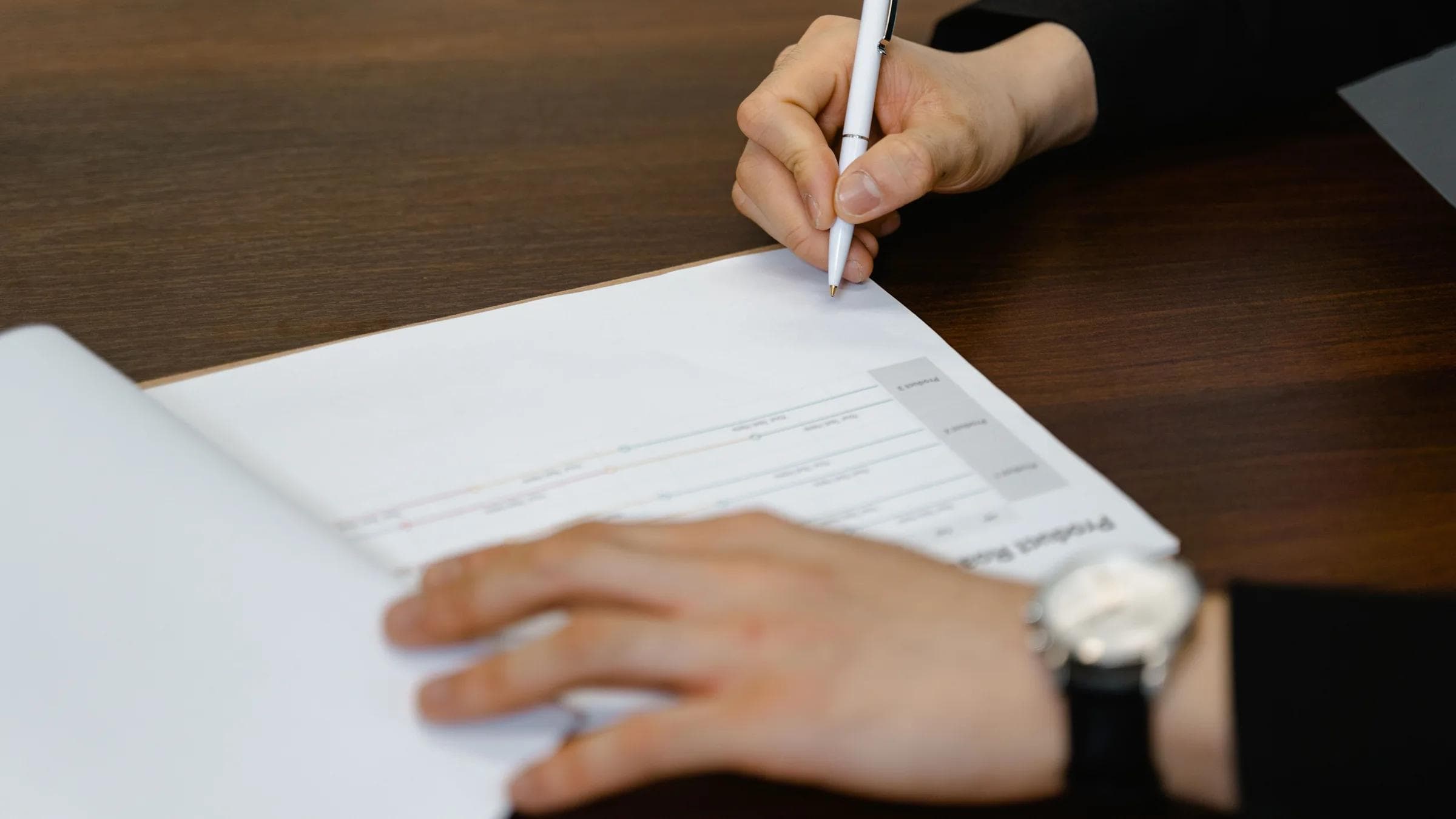 Businessperson writing on a document at a wooden desk, with focus on hands and pen.