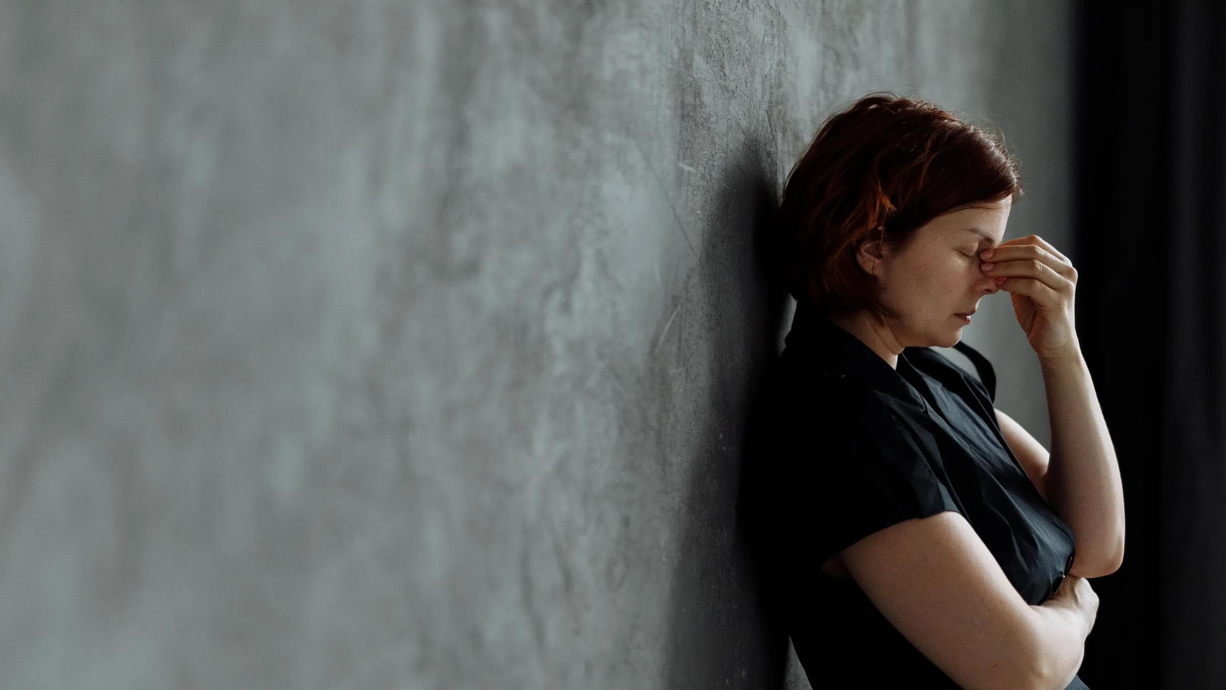 A woman in stress with eyes closed, leaning against a concrete wall indoors.
