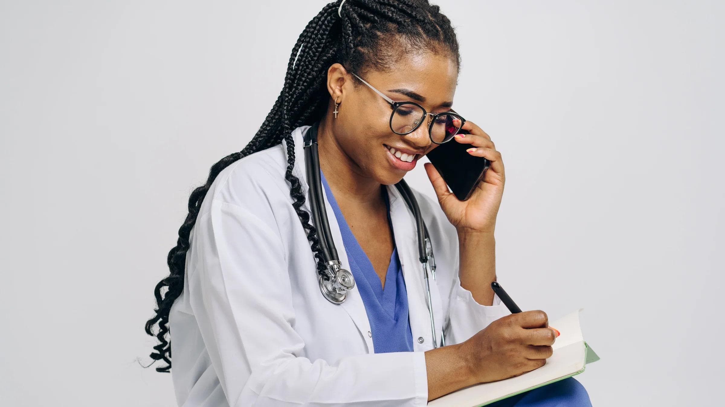 Female doctor smiling while talking on the phone and taking notes, wearing a stethoscope.