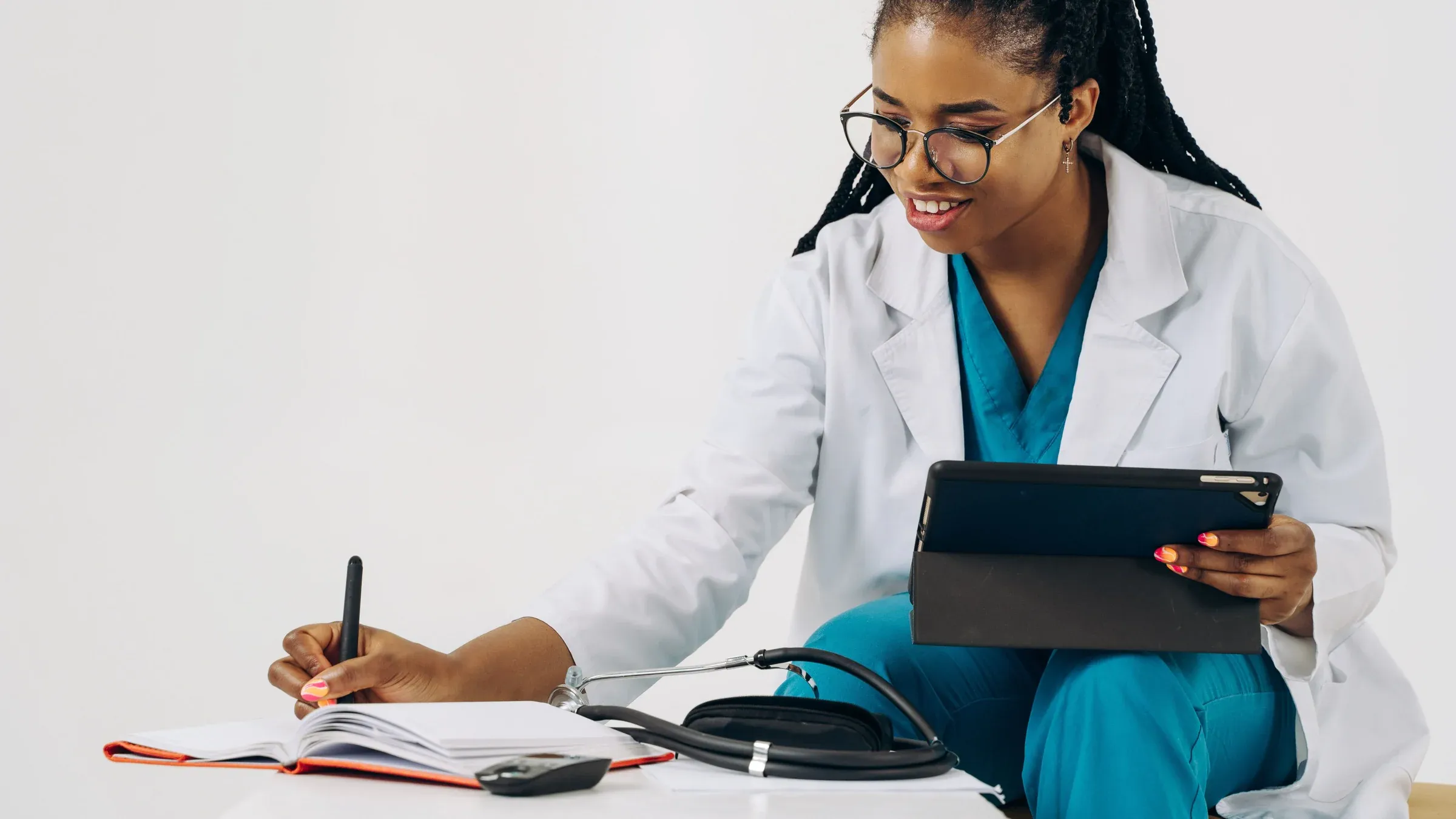 Smiling female doctor in scrubs using tablet and taking notes in a healthcare setting.