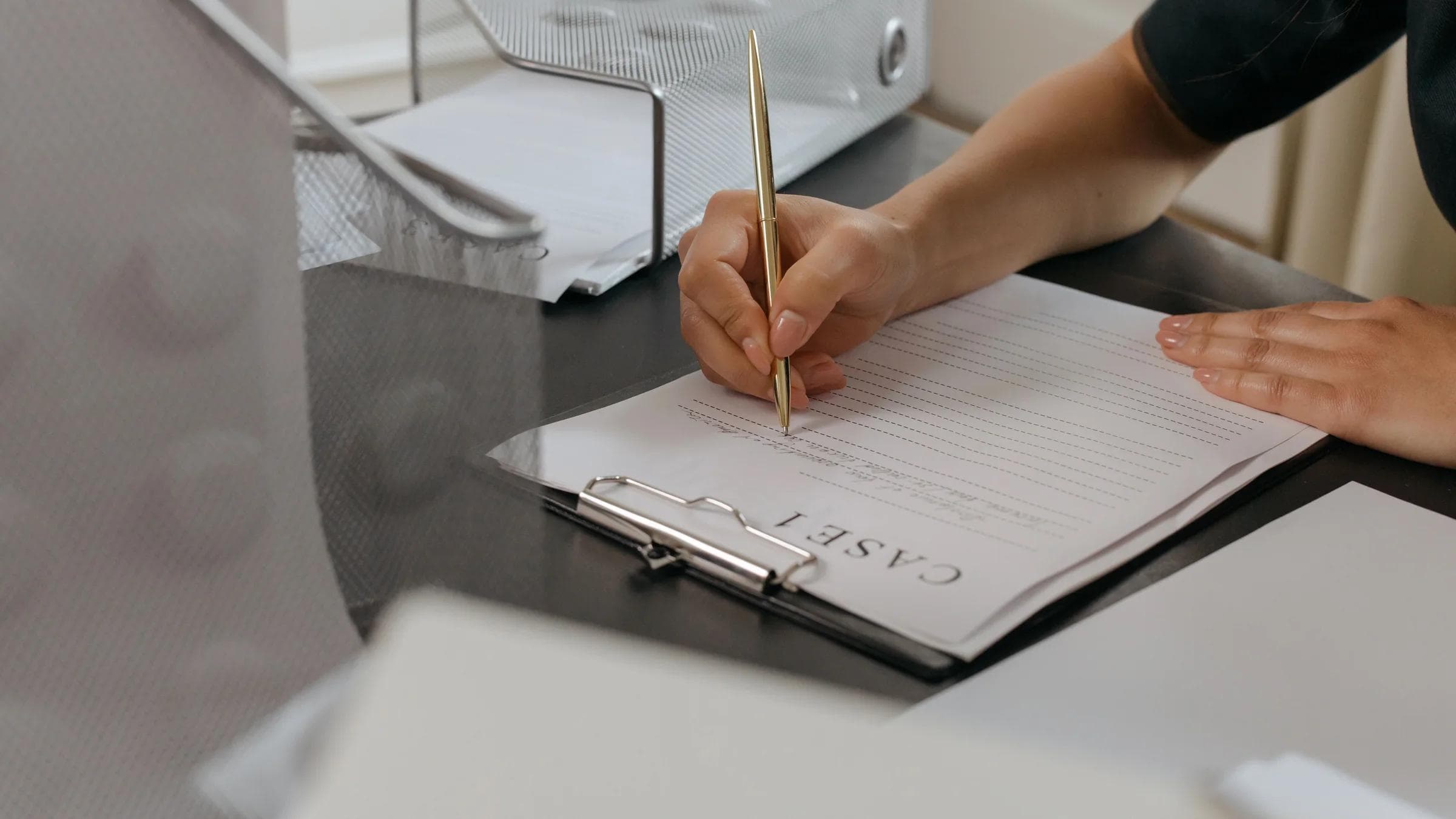 Close-up of a woman writing on documents with a gold pen in an office environment.