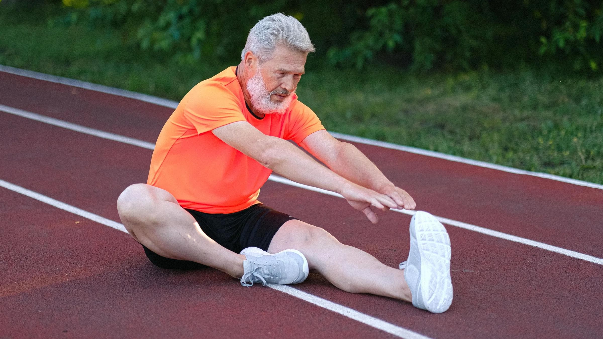 Full body of focused senior male sitting and stretching on racetrack in stadium near bushes with green leaves.