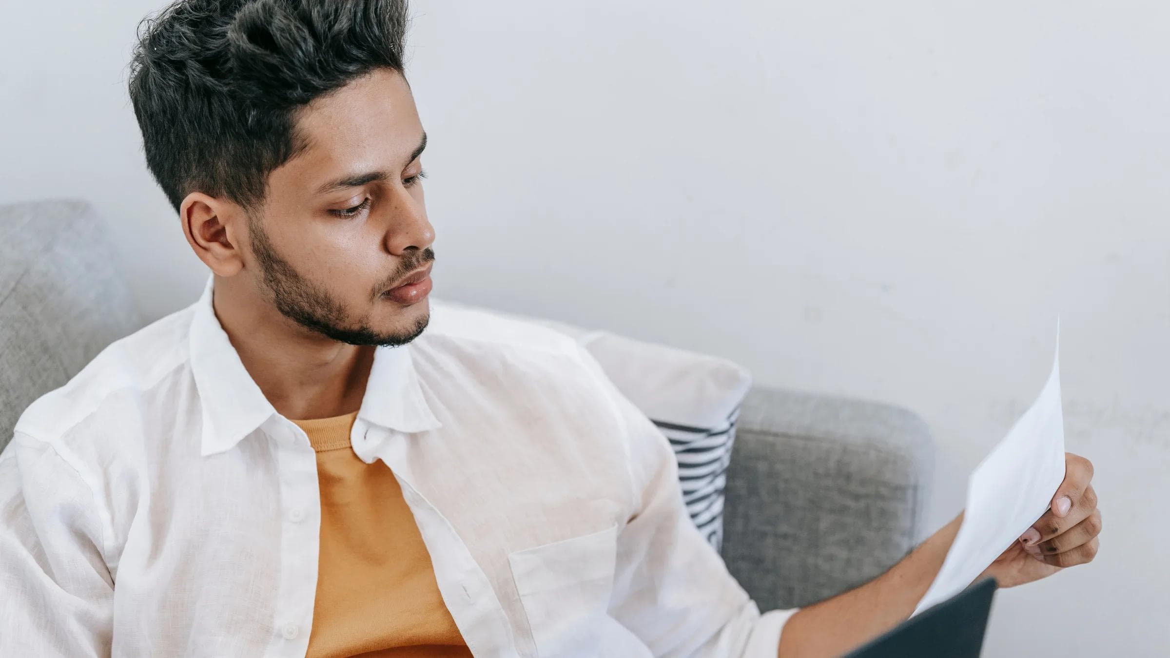 Concentrated young ethnic male wearing casual outfit reading document and browsing netbook while resting on cozy couch.