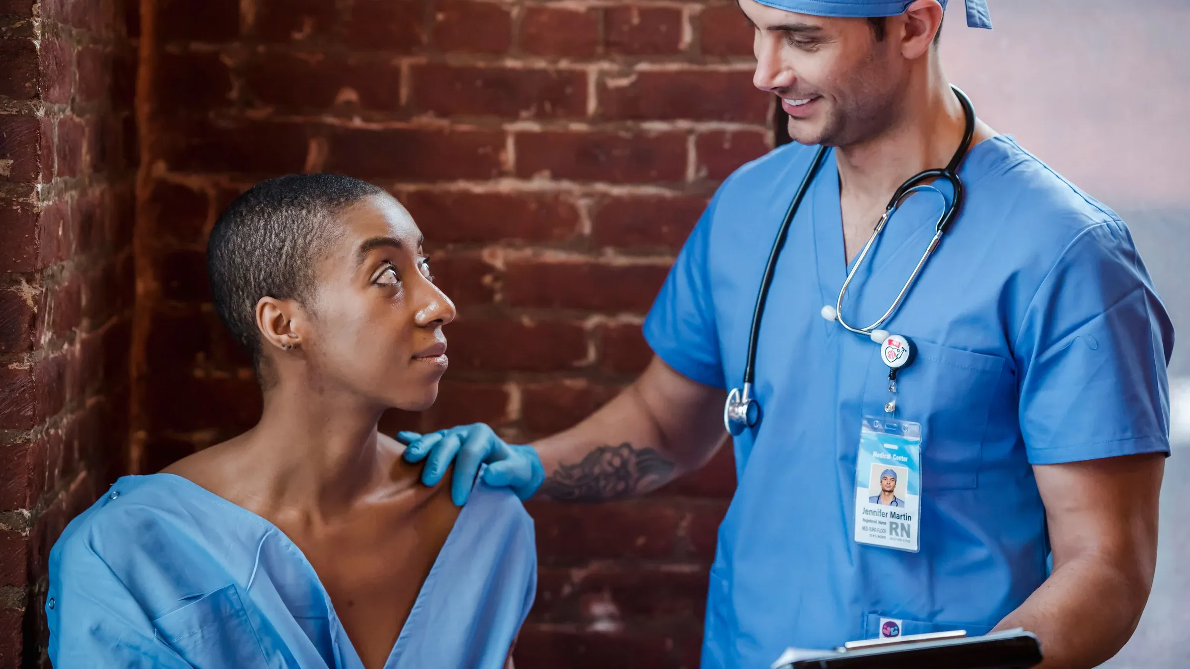 Positive doctor in uniform with stethoscope standing with clipboard and communicating with African American female person in short dark hair in hallway of clinic in daytime.
