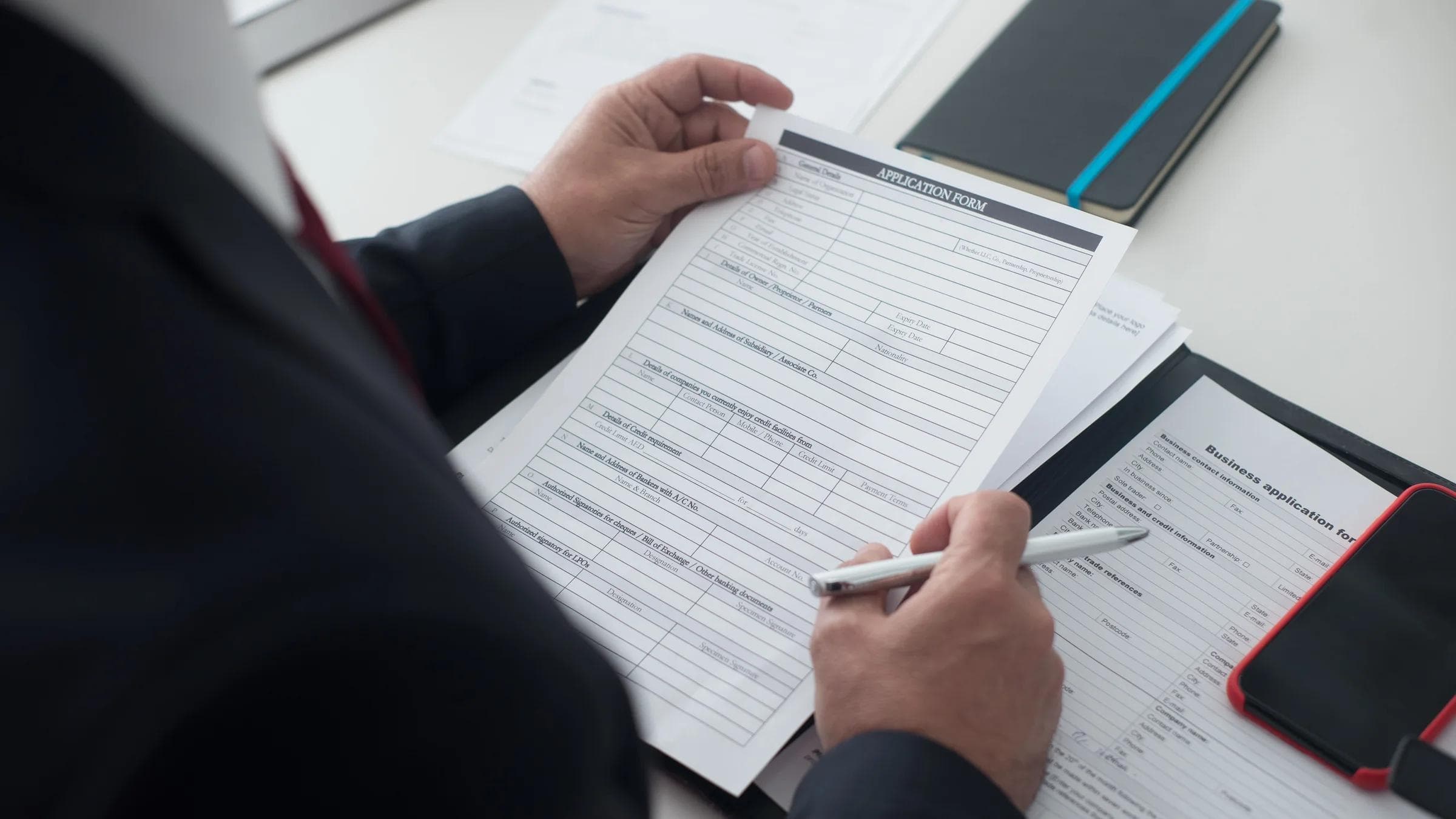 A businessman in a black suit reviews a printed application form with a pen on his desk.