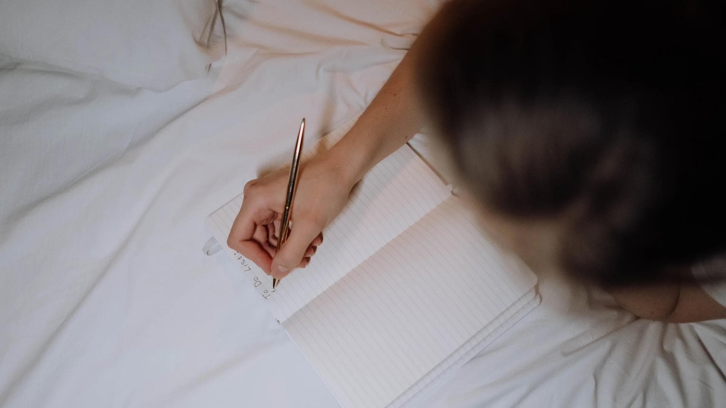 A person writing a to-do list in a notebook while sitting on a bed with white linen.