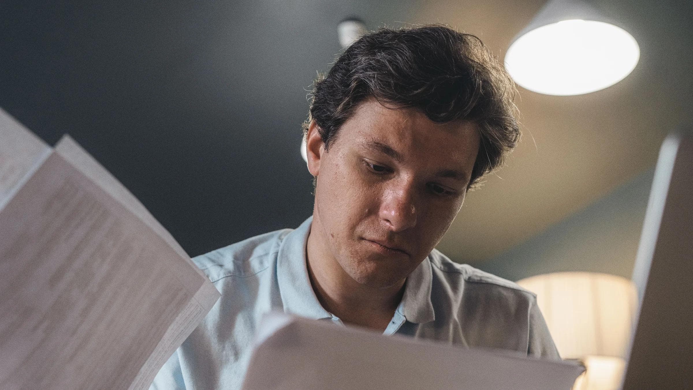 Serious man wearing a light blue shirt intently reading printed papers indoors under warm lighting.