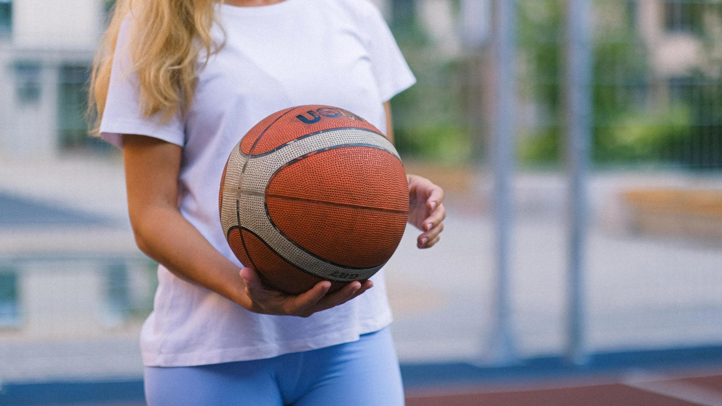Crop unrecognizable woman in activewear on basketball court with ball near fence in daylight.