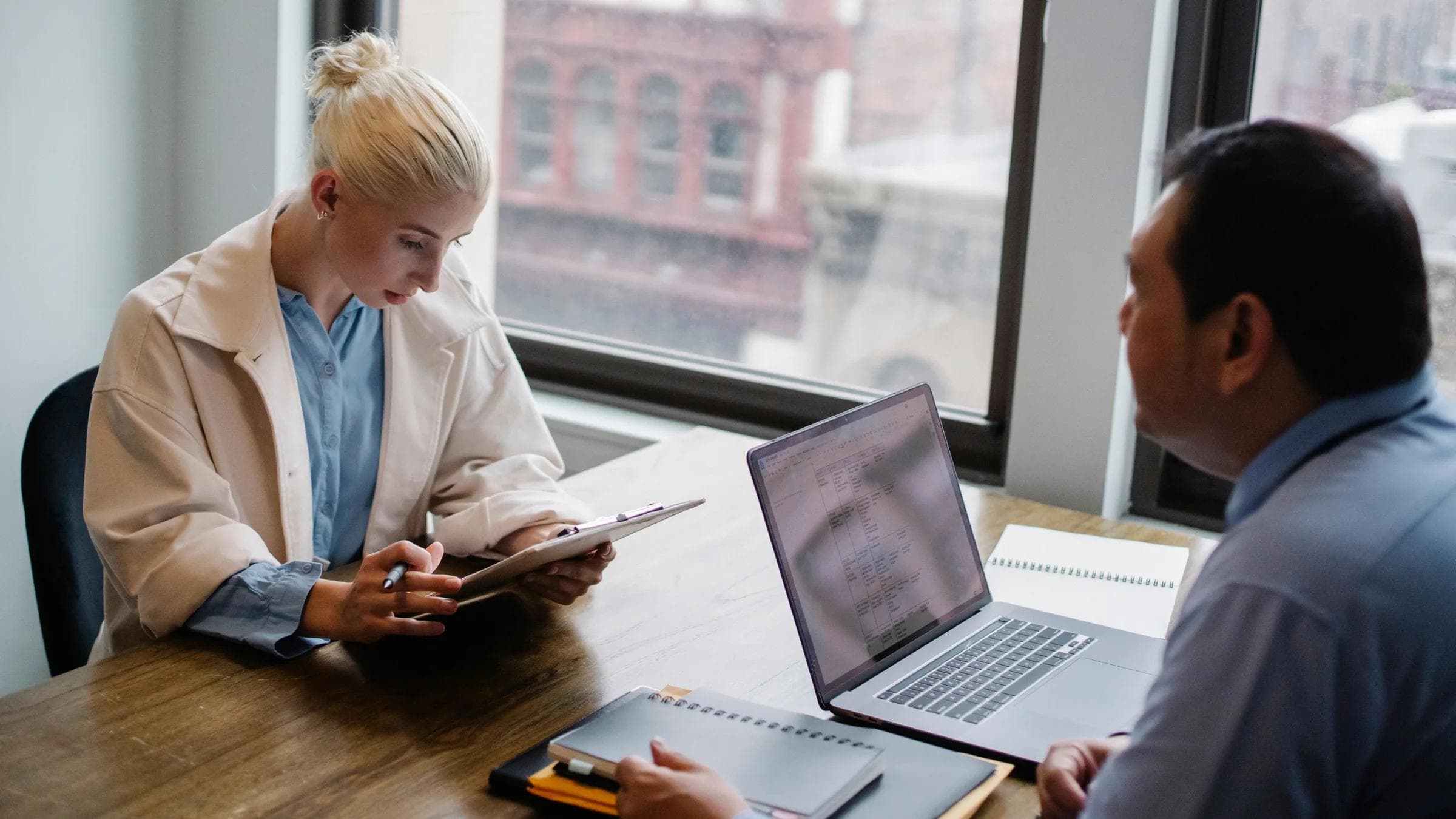 From above of young female worker wearing modern coat analyzing details of project with colleague while sitting together at wooden desk with laptop and documents.