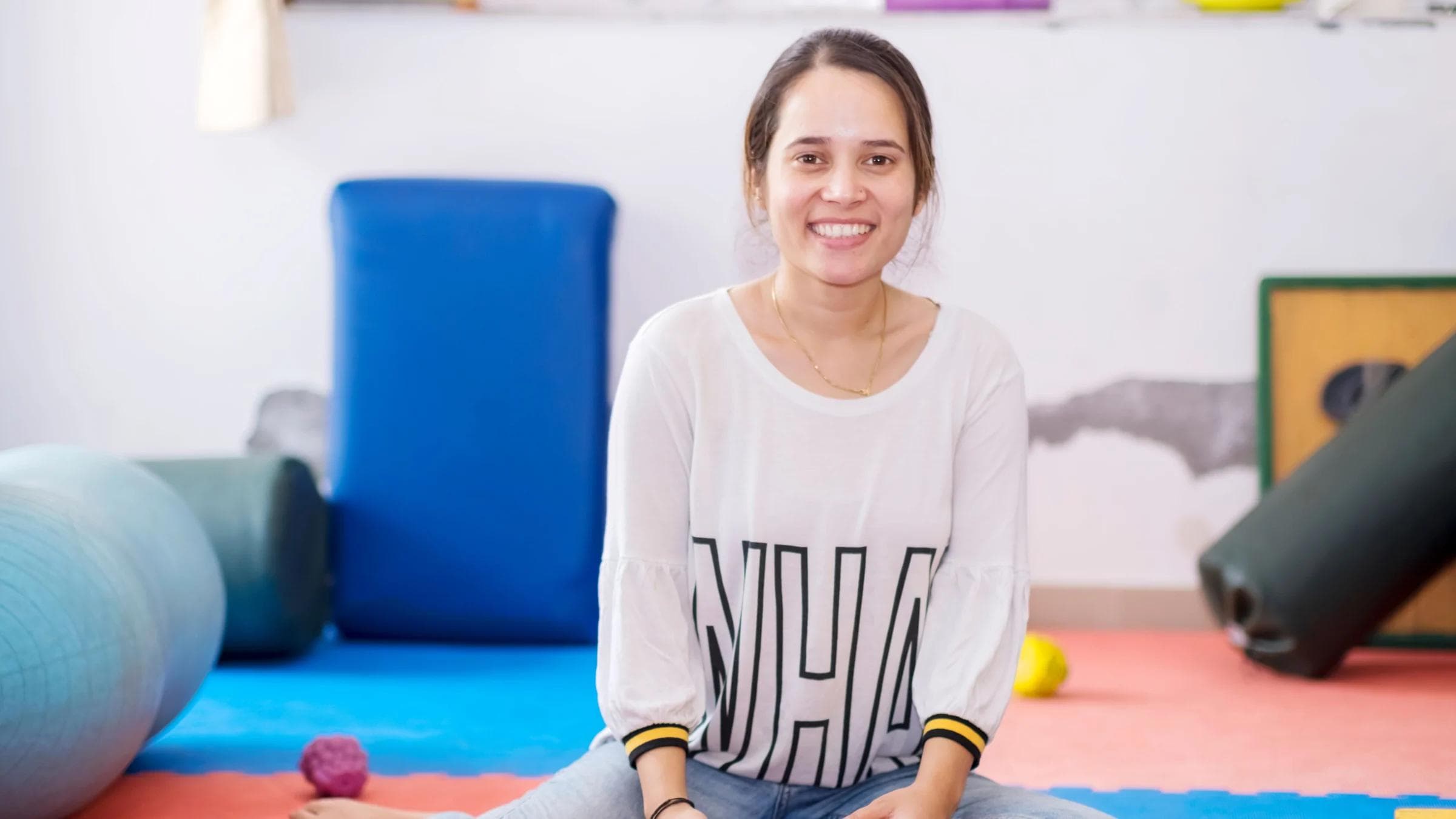 Smiling woman holding therapy balls, seated in a brightly colored fitness studio.