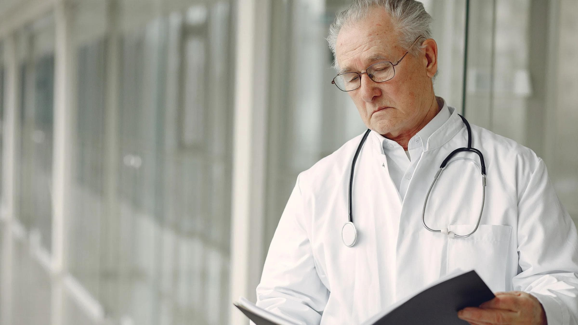 Crop concentrated professional senior male physician in white robe with stethoscope standing in modern hospital hallway and reading medical case records in daylight.