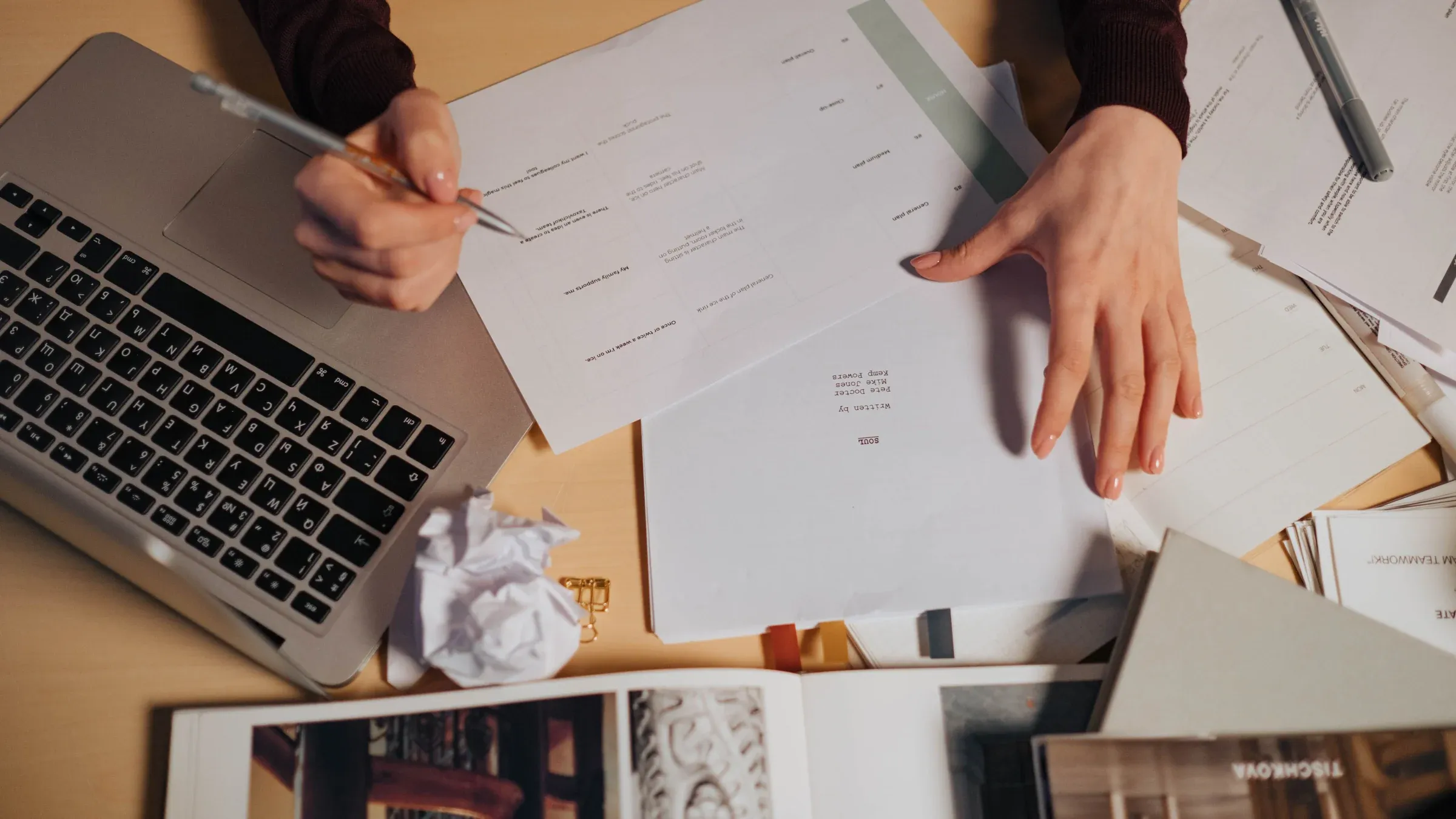 A person taking notes and working on a laptop surrounded by documents on a desk.