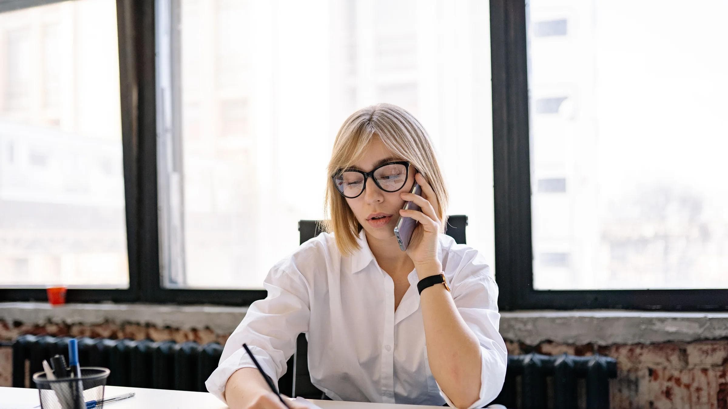 A woman in an office setting multitasking with a phone call and taking notes.