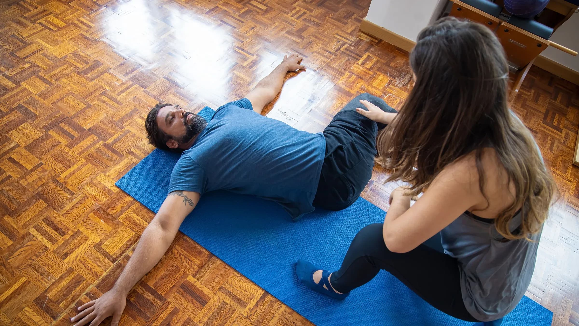 A man and woman engaged in a rehabilitation exercise on a yoga mat indoors.