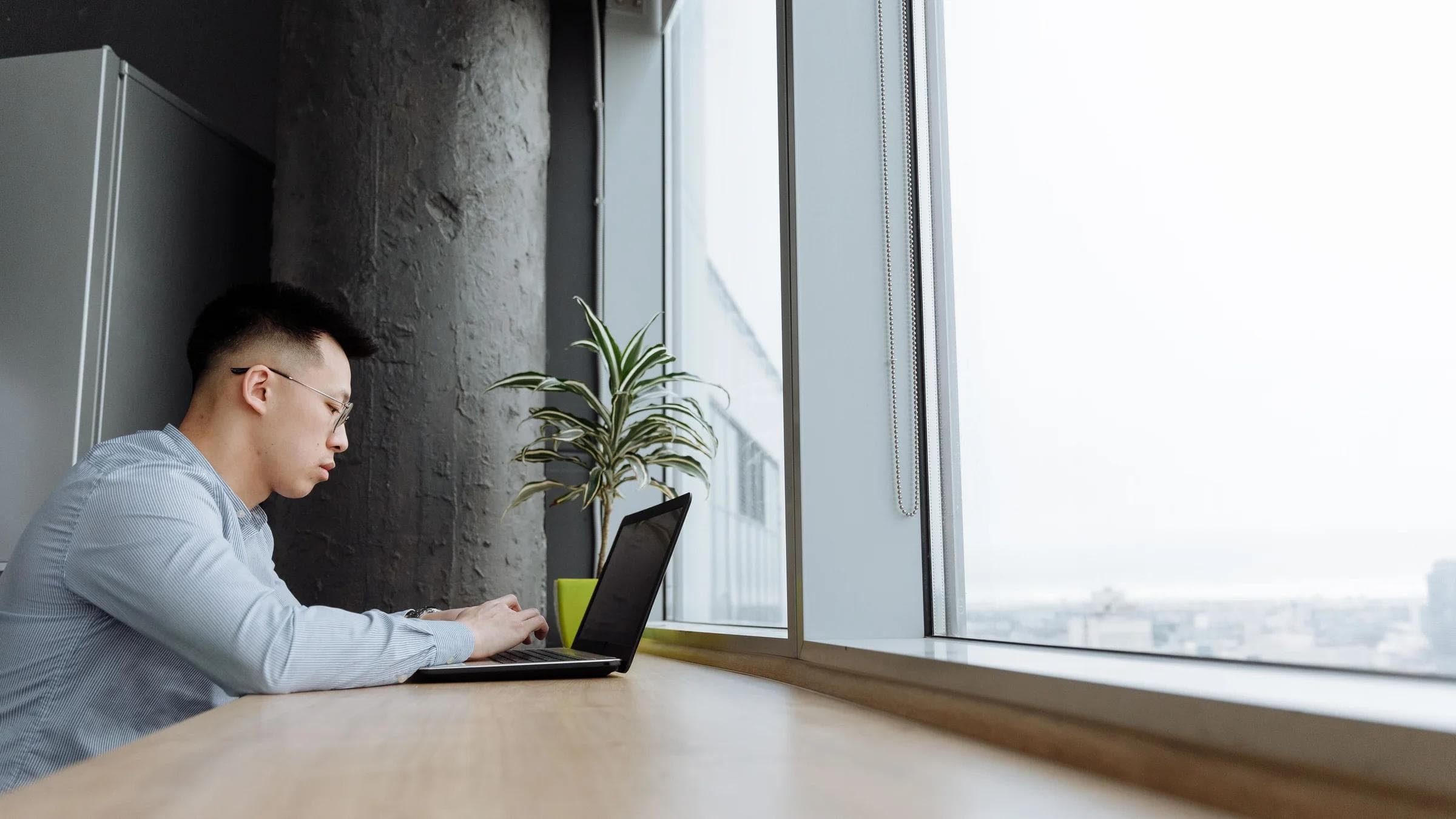 Asian man engaged in work on a laptop in a bright, modern office setting with large windows.