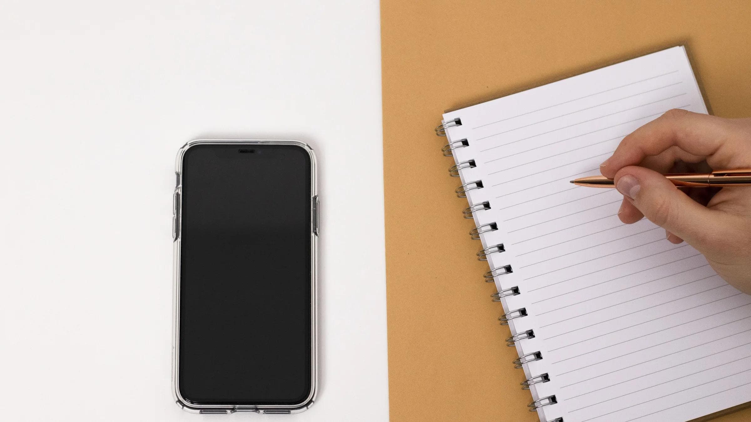 Minimalist top view of a workspace setup featuring a smartphone and notebook with a hand holding a pen.