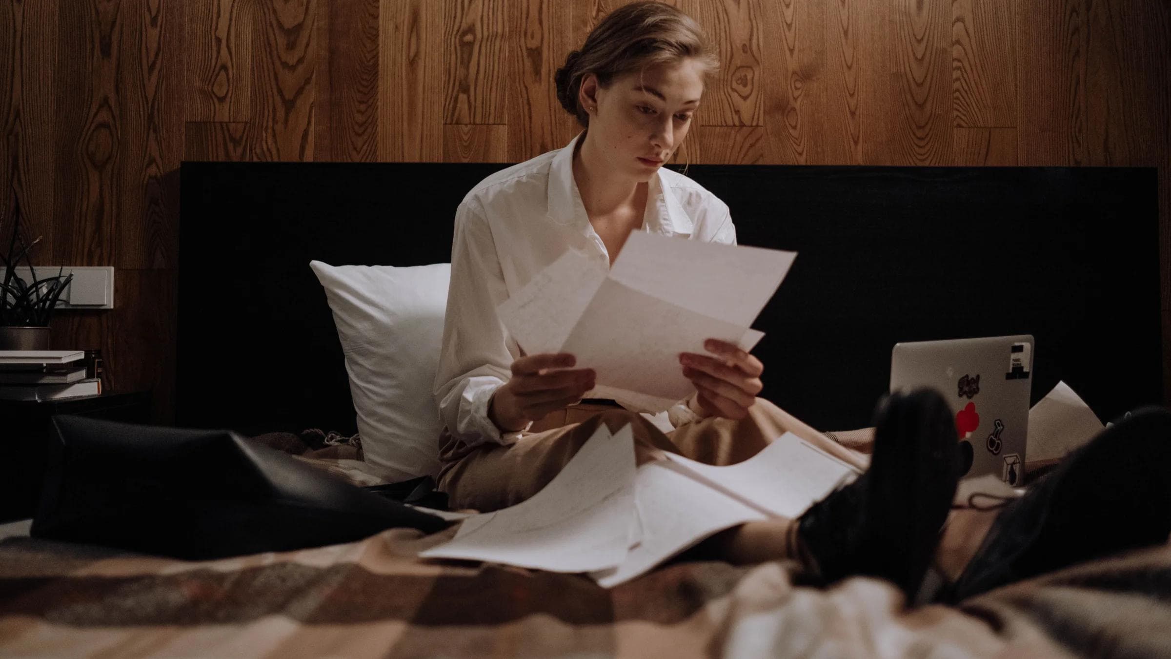 Woman sitting on bed, working from home with laptop and paperwork, focused and thoughtful.