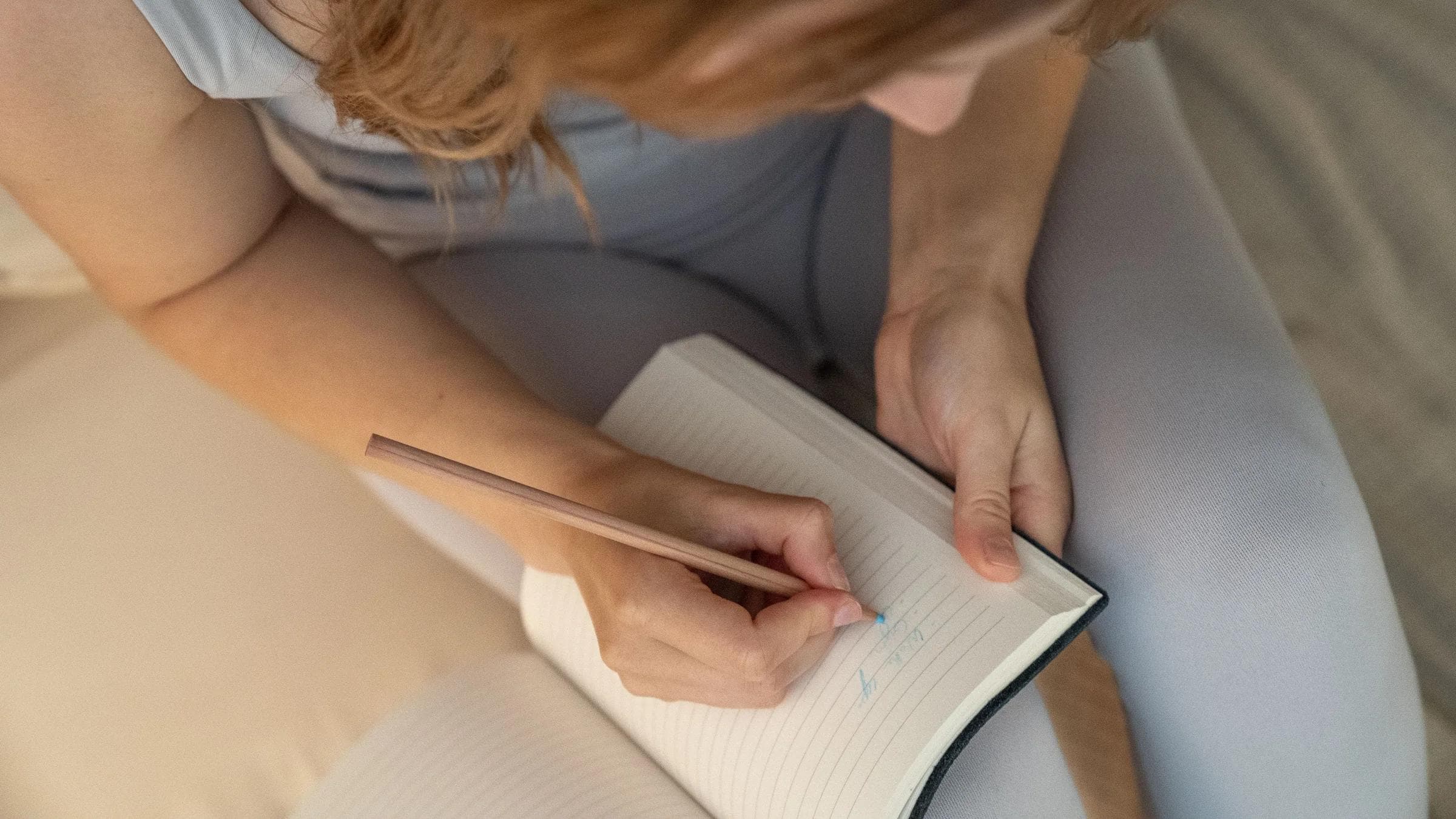An adult woman writes in a notebook with a pencil, viewed from above.