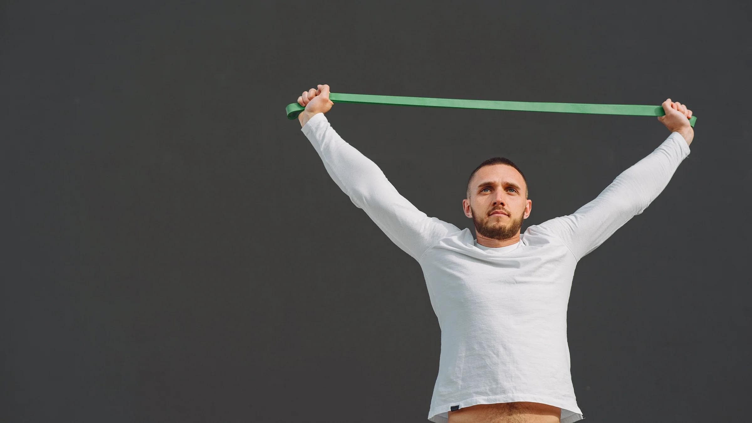 Adult male in sportswear exercising with a green resistance band against a gray backdrop.