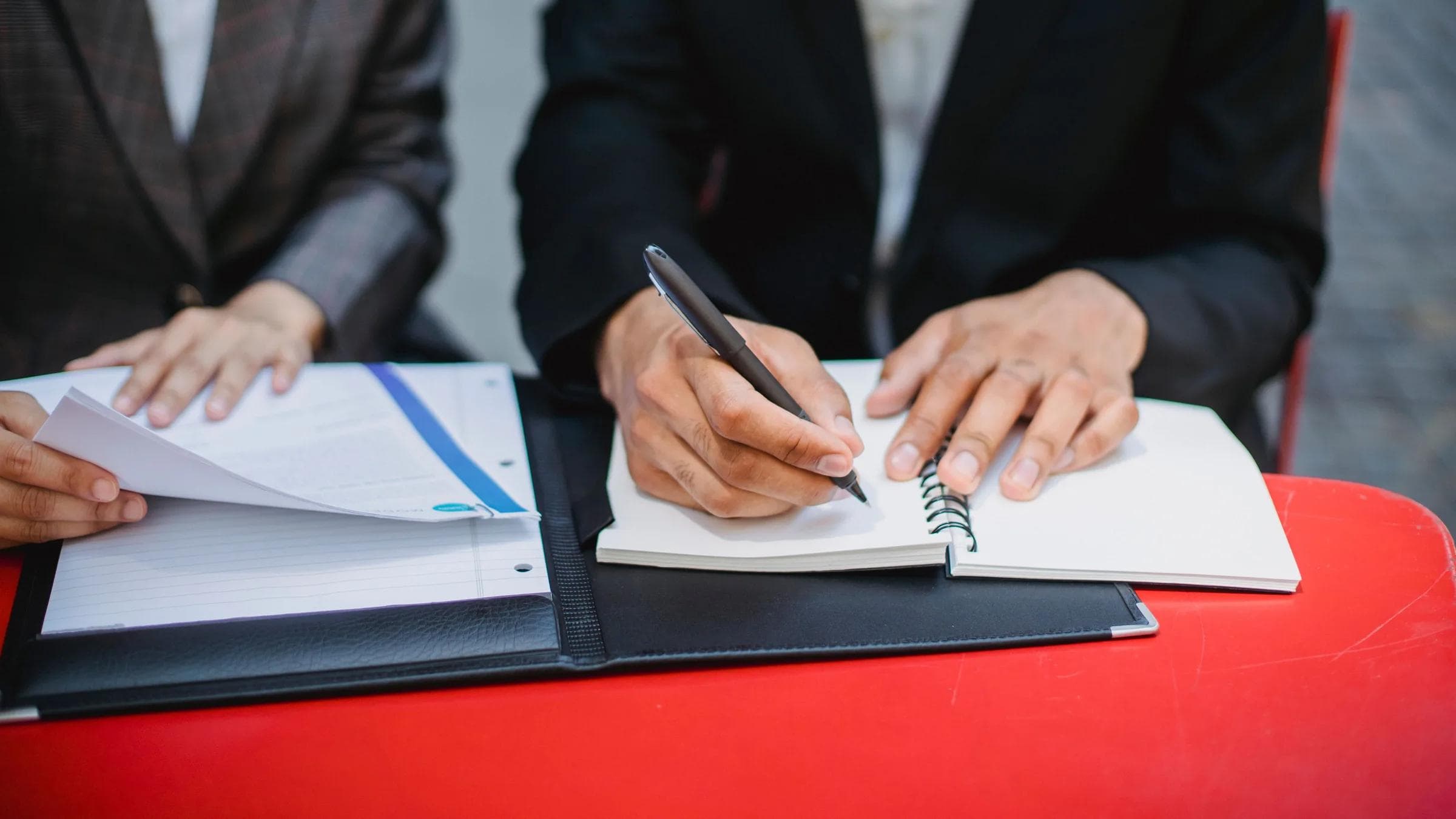 Two professionals in suits writing notes and reviewing documents at an outdoor meeting table.