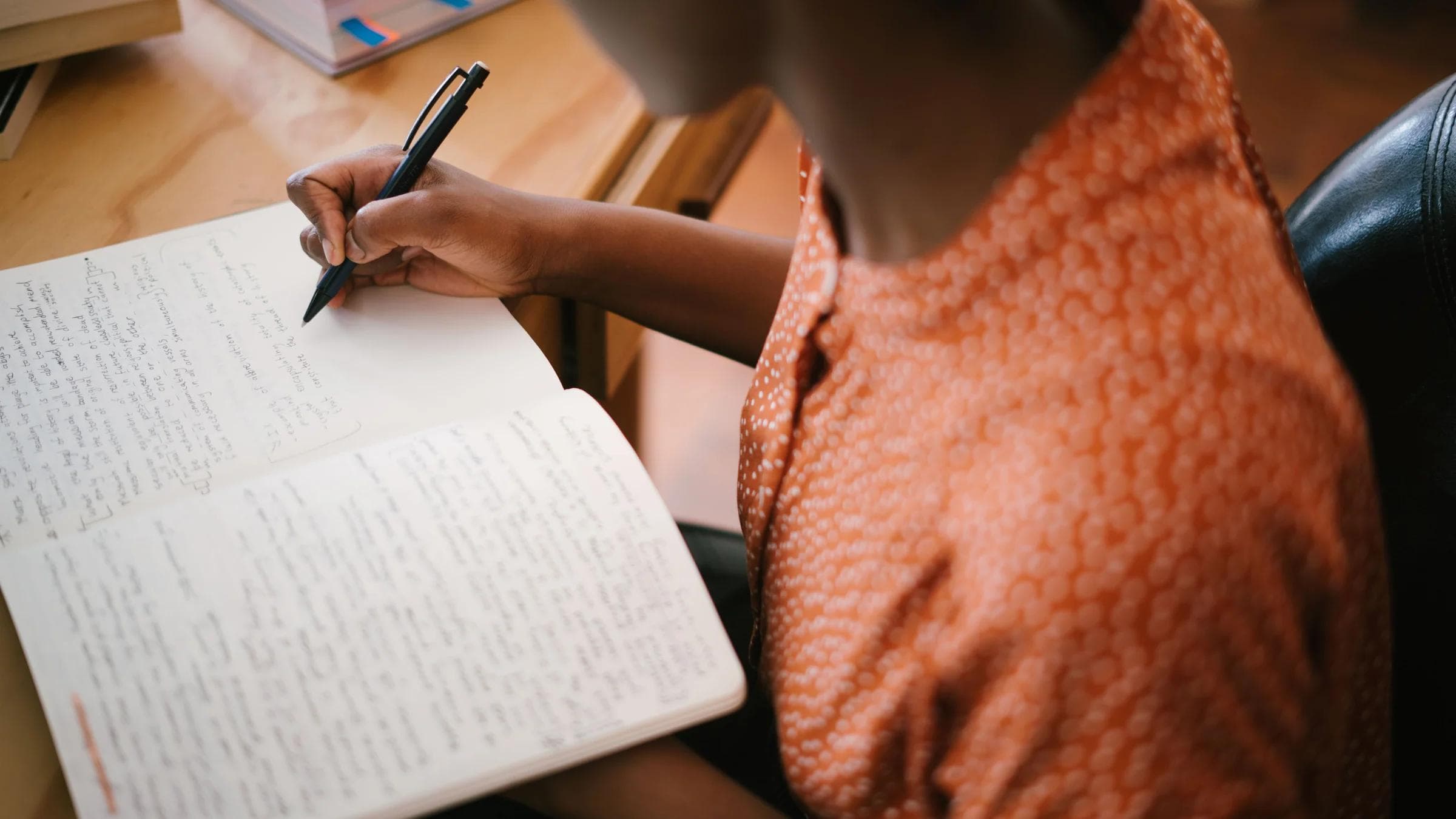 Focused black woman writing in a notebook with stacked books on a desk, illustrating learning.