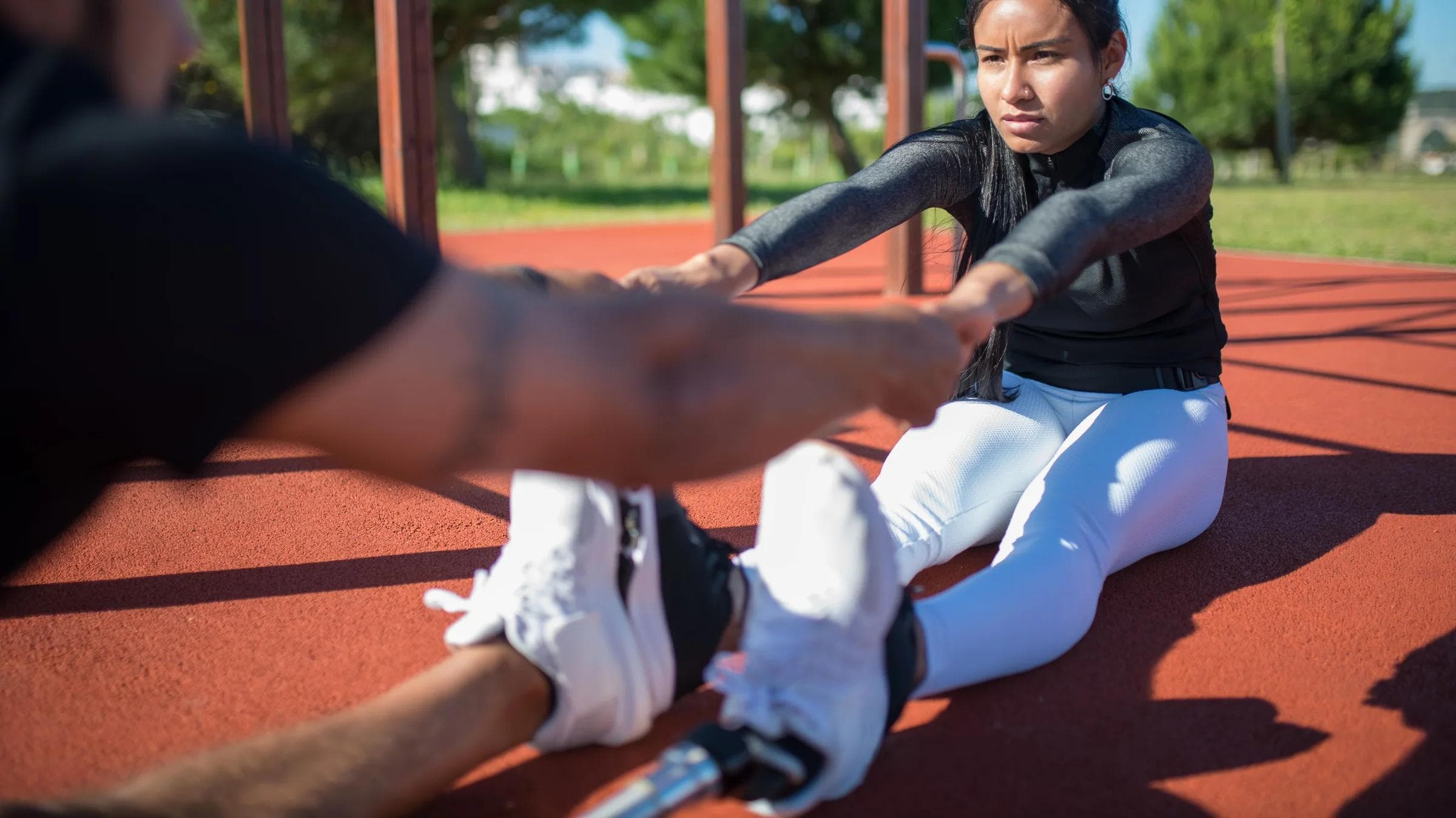 A woman performing a stretching exercise in a sunny park with a coach's assistance.