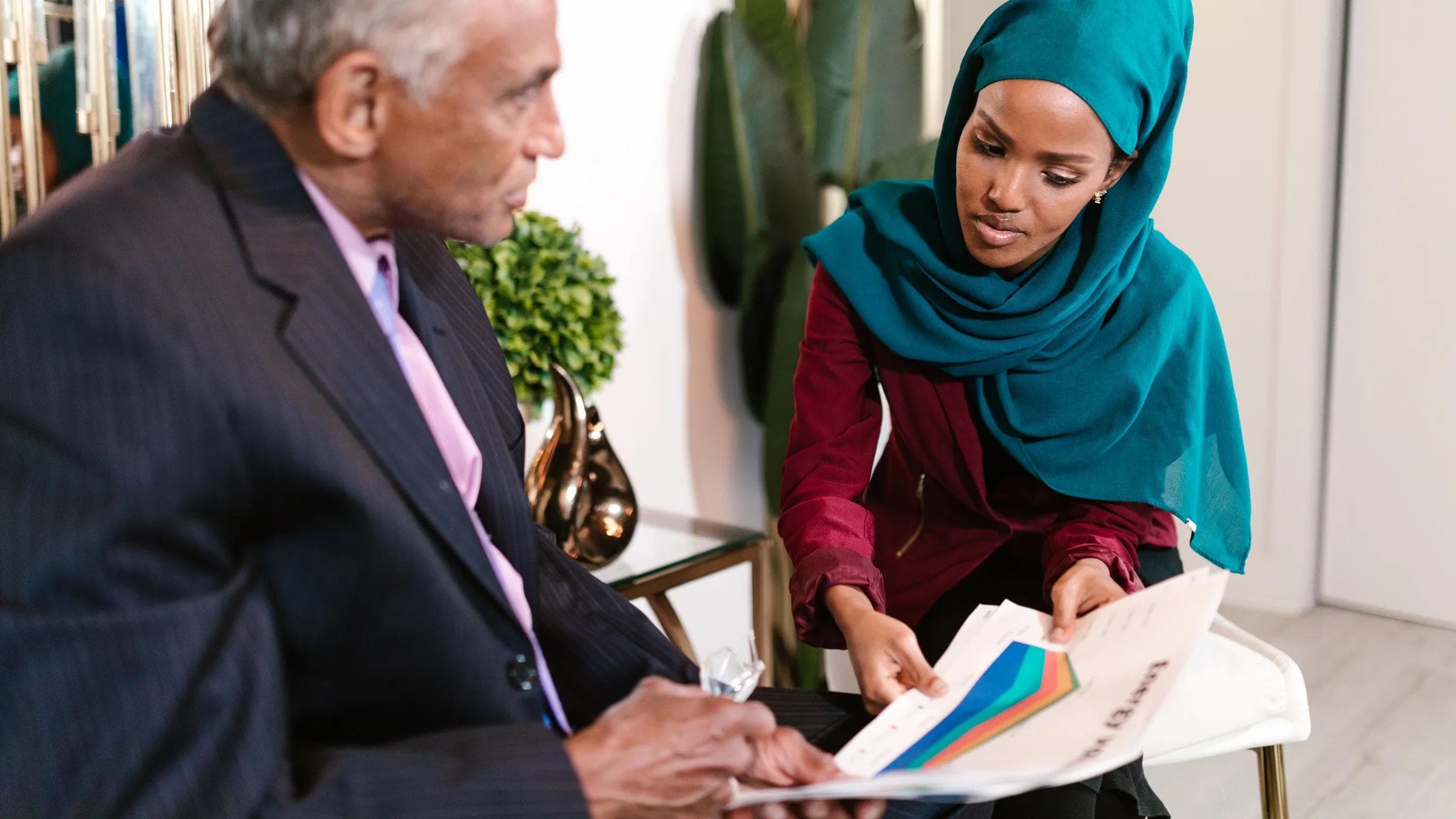 A man and woman in business attire discuss documents in a modern office setting.
