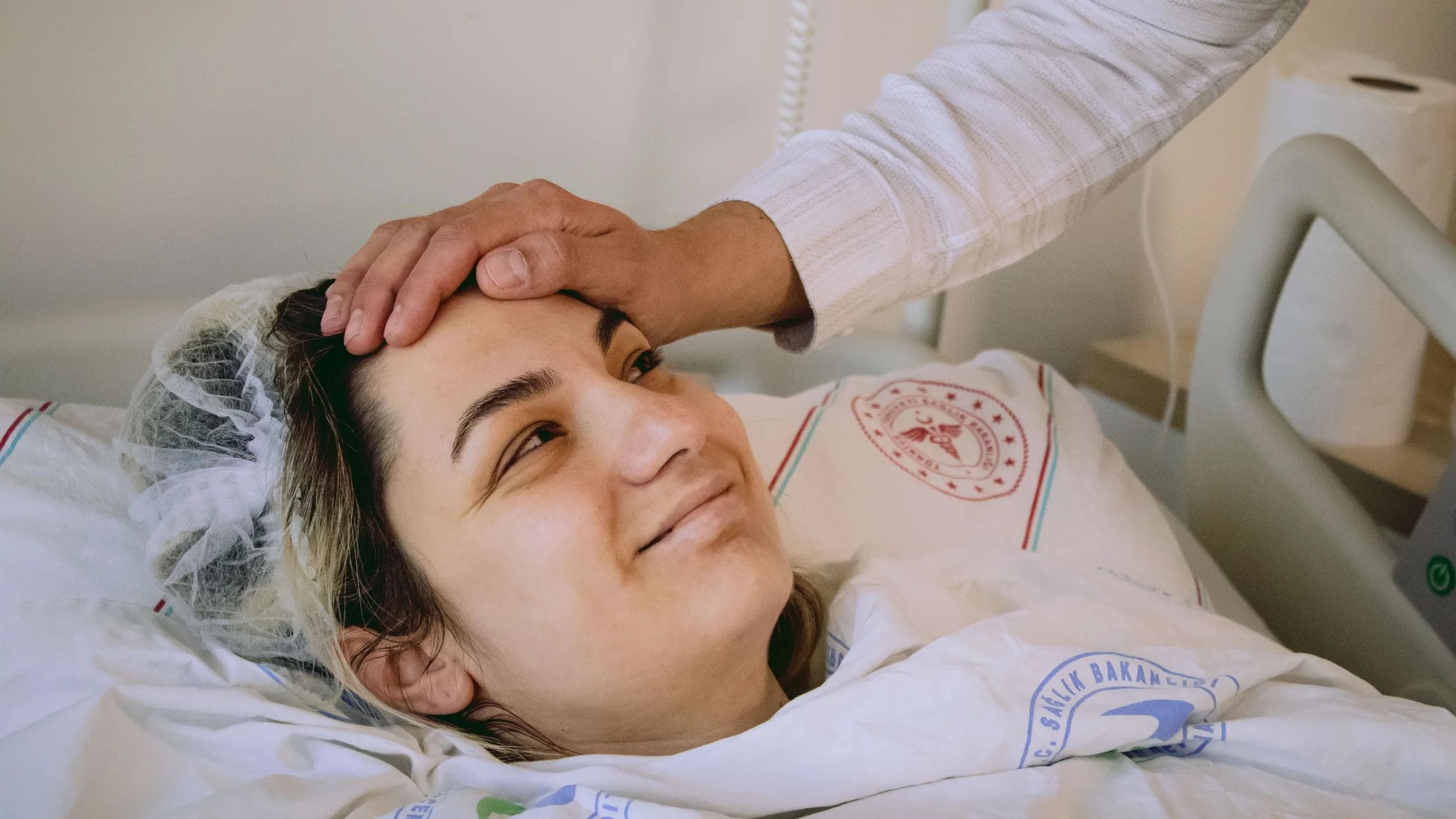 Smiling woman in hospital bed with supportive hand on forehead, conveying warmth and care.