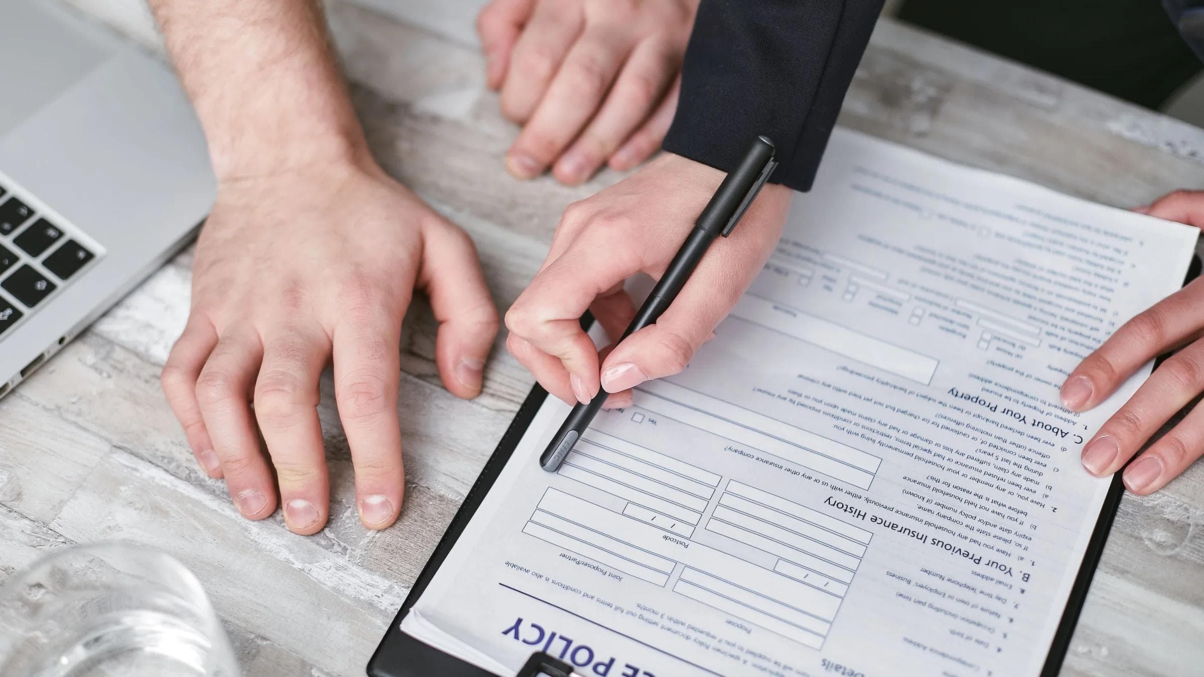 Two adults sign an insurance policy document at a wooden desk.