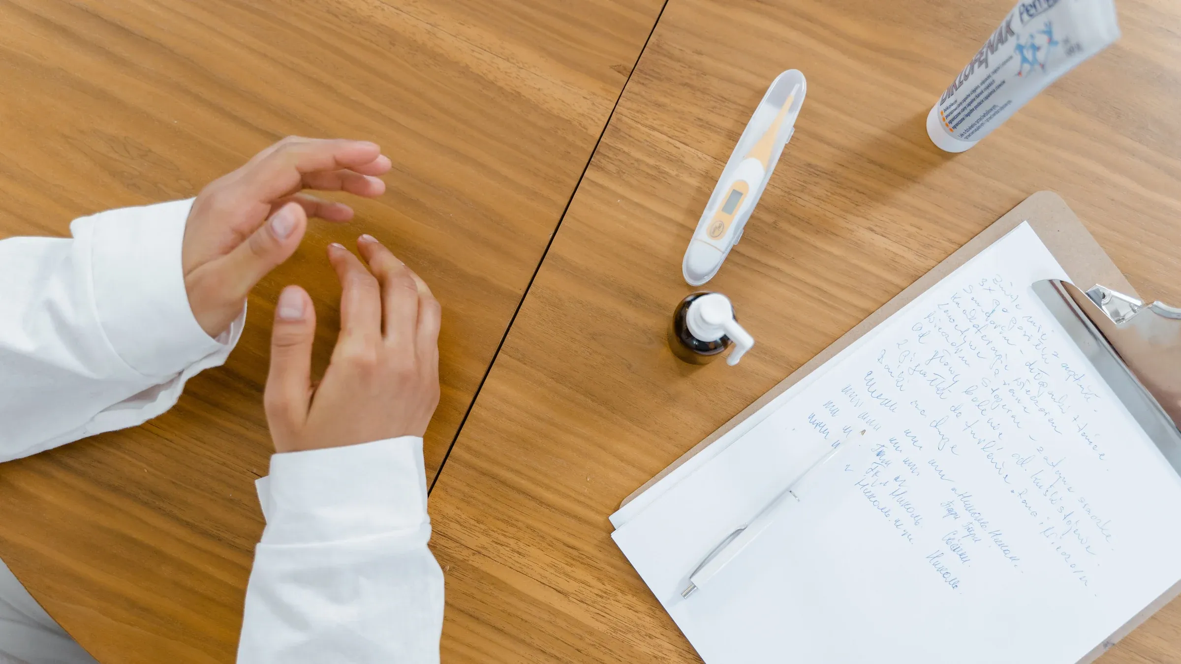 Top-down view of a doctor's desk featuring notes, thermometer, and hands in a healthcare setting.