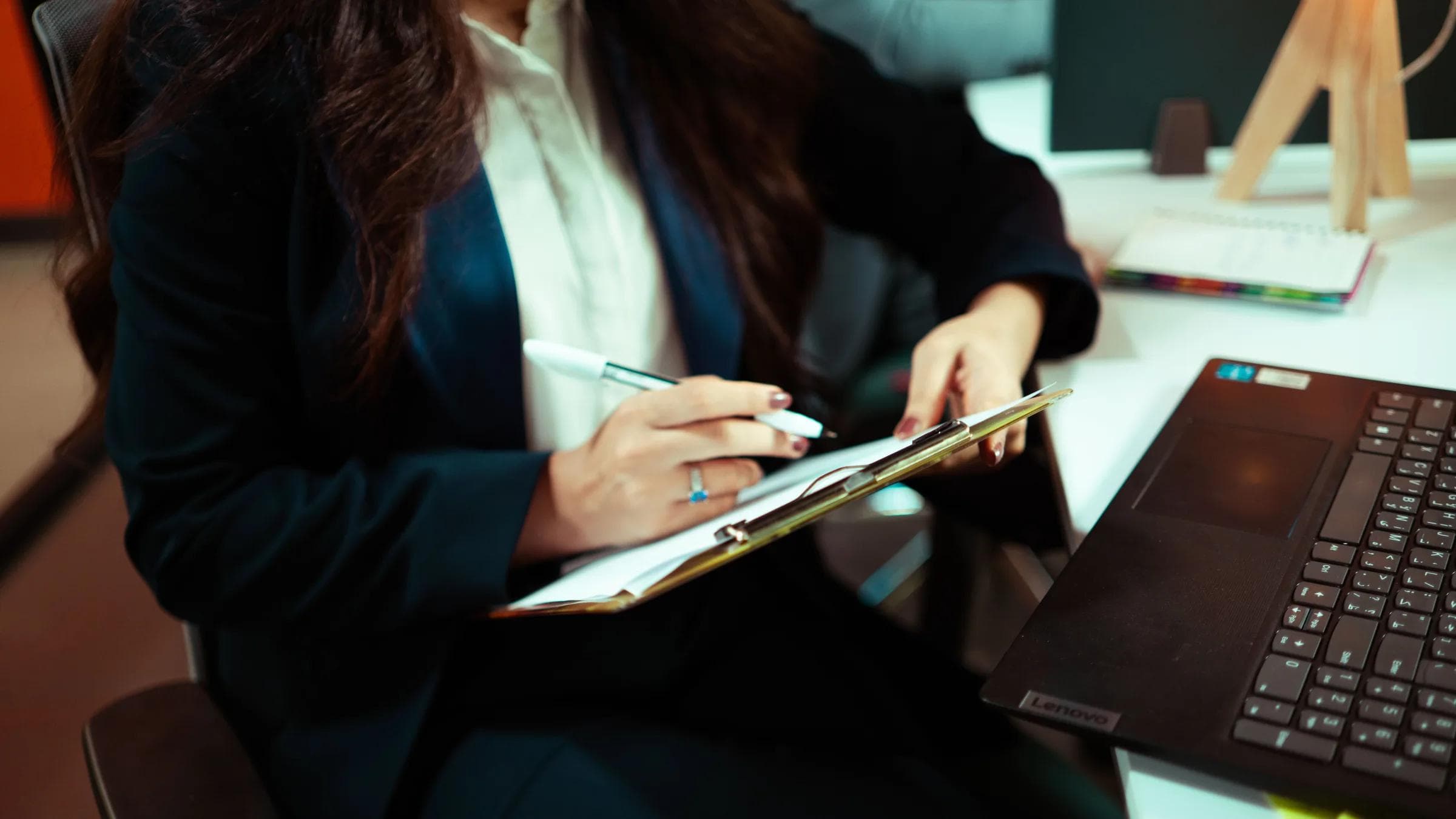 A focused businesswoman writing on a clipboard with a laptop nearby in an office environment.