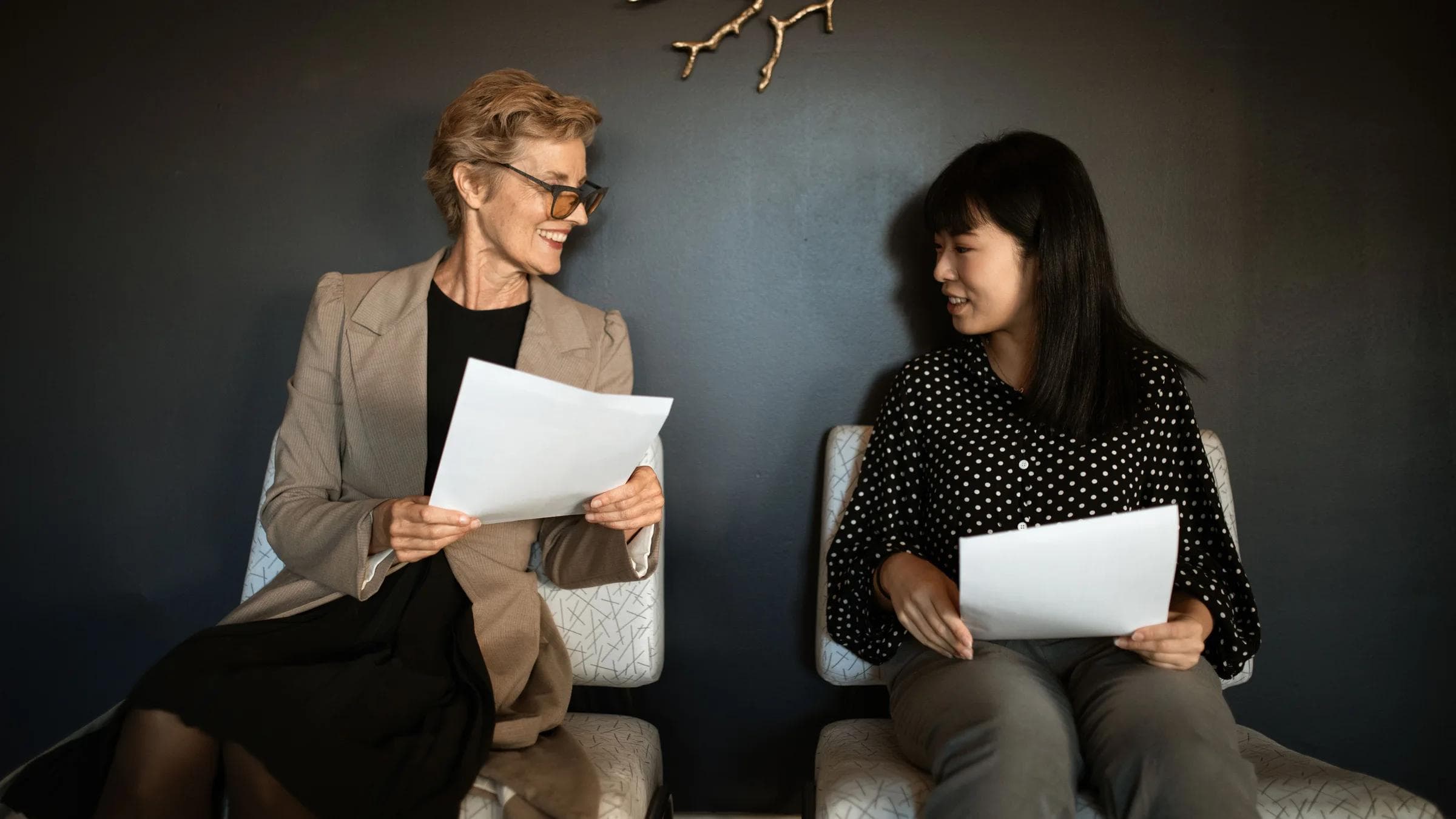 Two women enjoying a friendly discussion indoors holding papers. Bright and relaxed atmosphere.