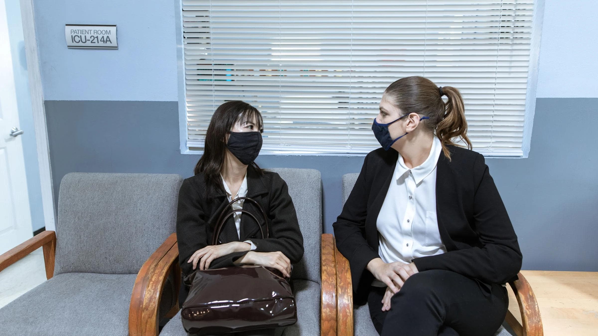 Two women wearing face masks sit in a hospital waiting area, reflecting the new normal during the pandemic.