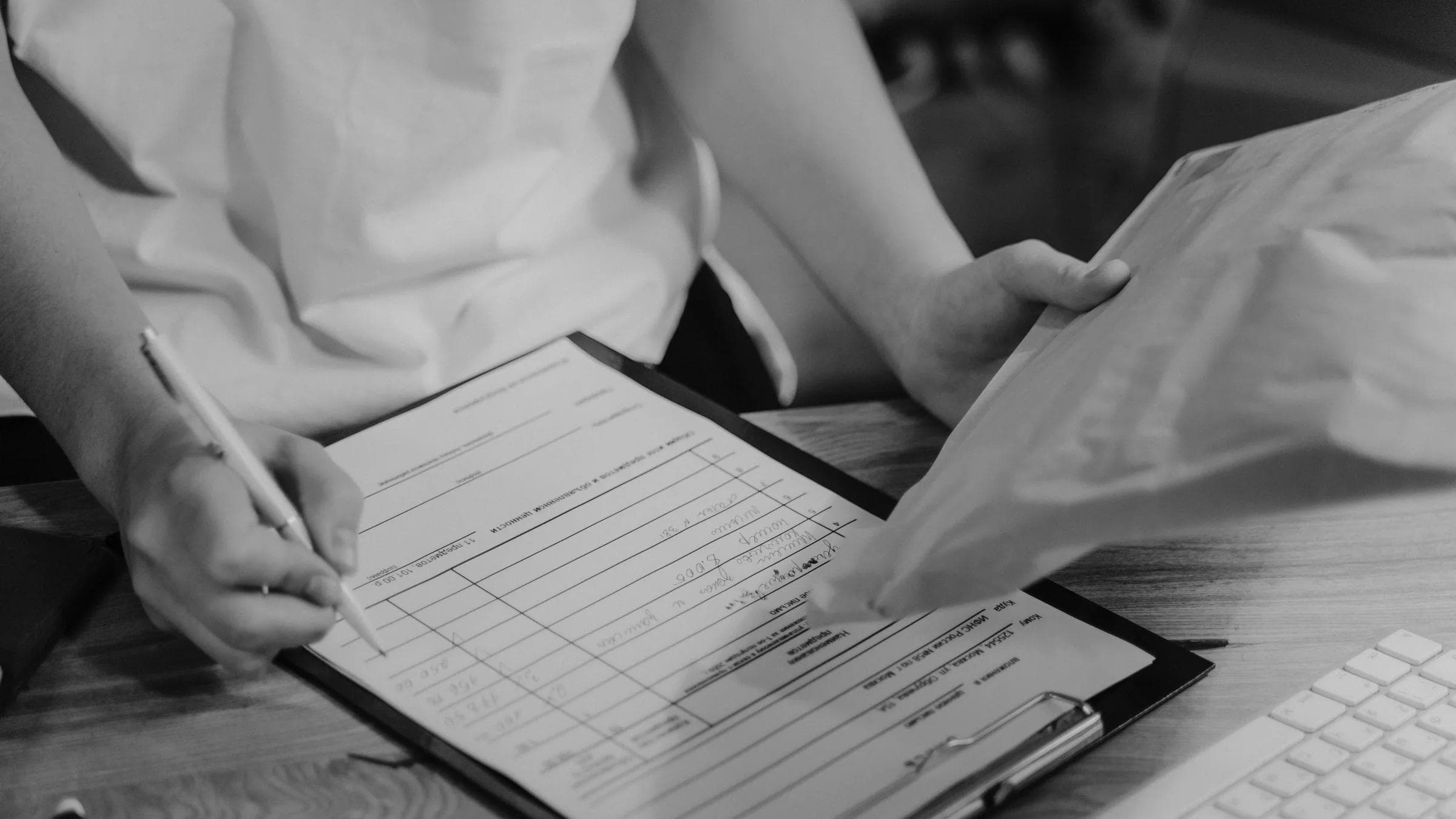 Black-and-white photo of a person writing on a clipboard with a keyboard visible.