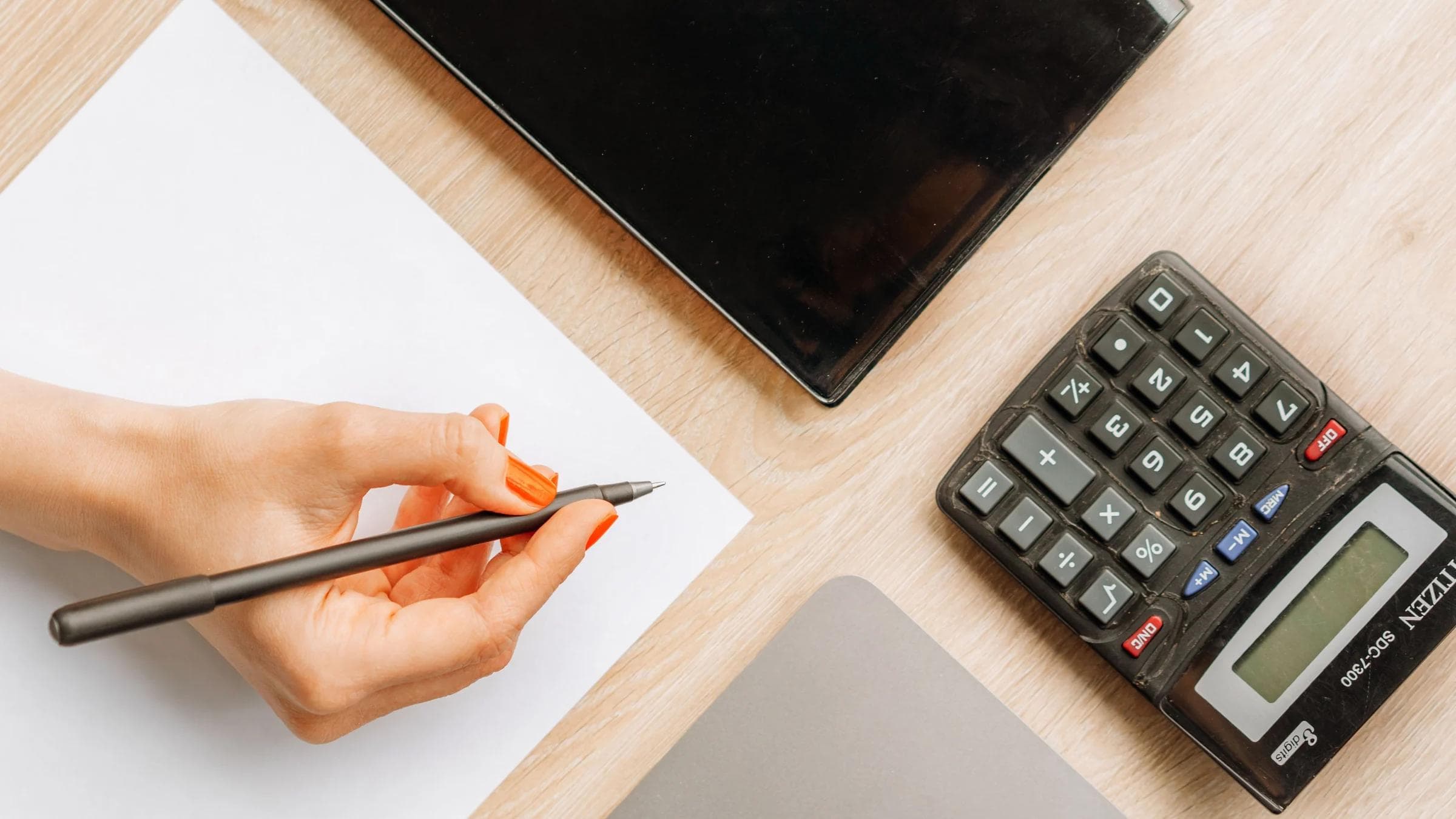 Top view of a minimalist office desk with a hand writing, calculator, and electronic device.