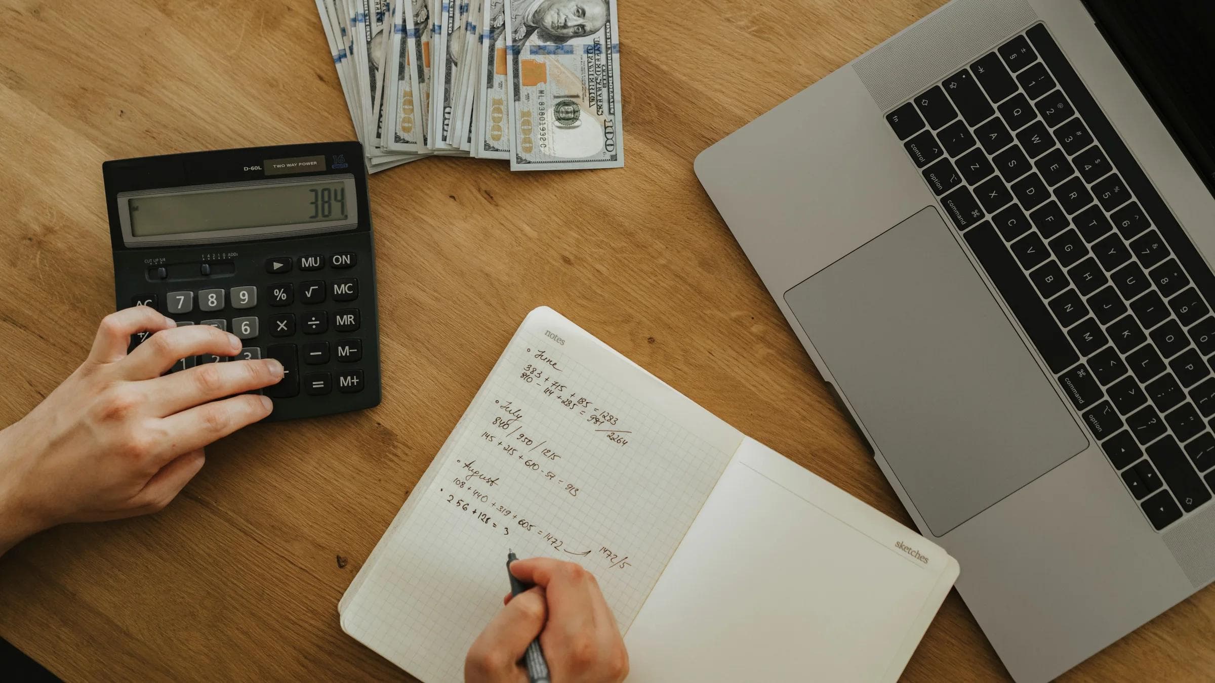 Hands using calculator and notebook, planning finances, with cash and laptop on wooden table.