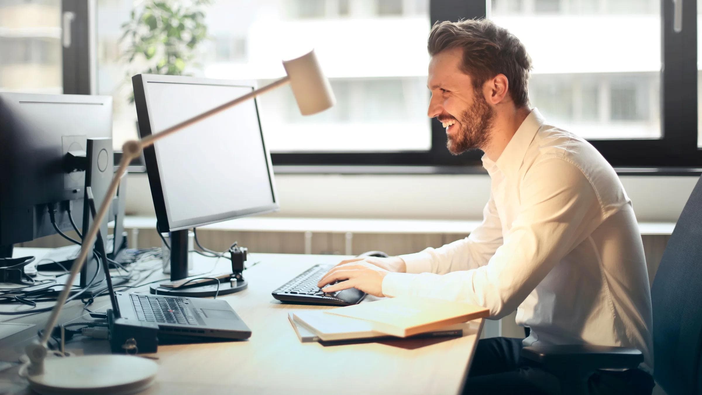 A man smiling while working at an office desk with a computer and natural daylight streaming in through large windows.