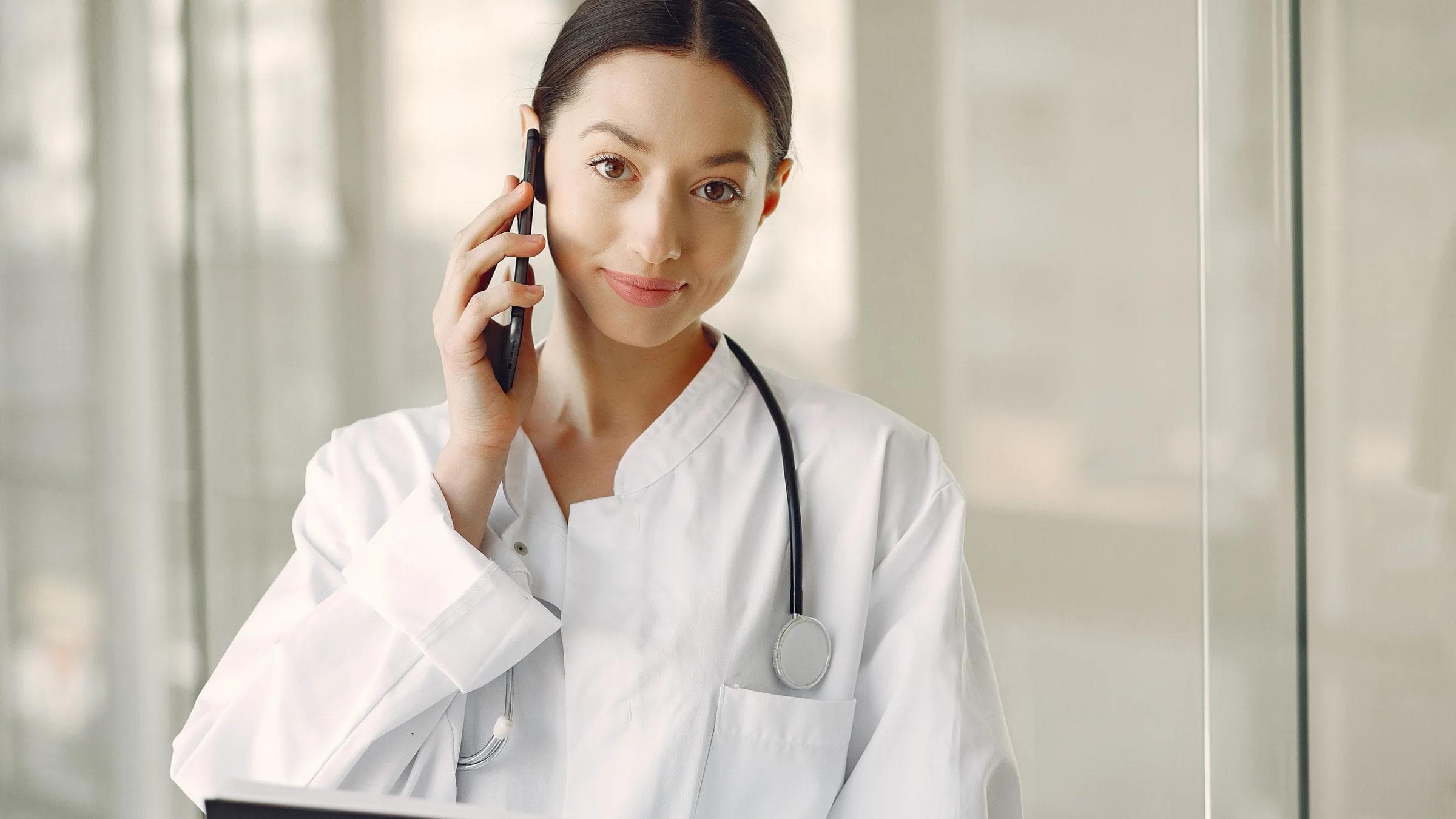 Crop ethnic female doctor in medical uniform with stethoscope and notebook talking on cellphone while standing in clinic corridor.