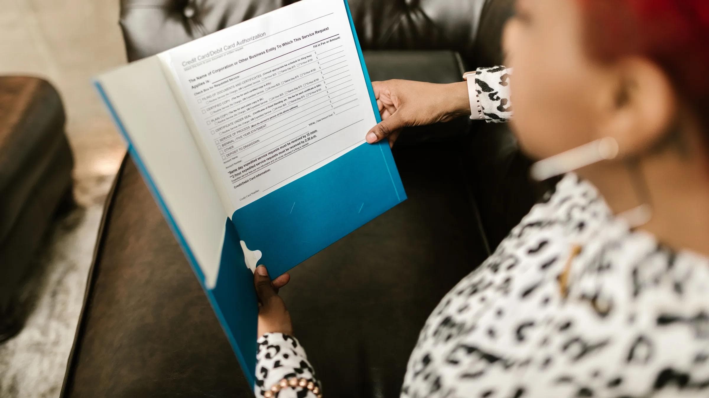 A woman sits on a couch reading a document in a blue folder.