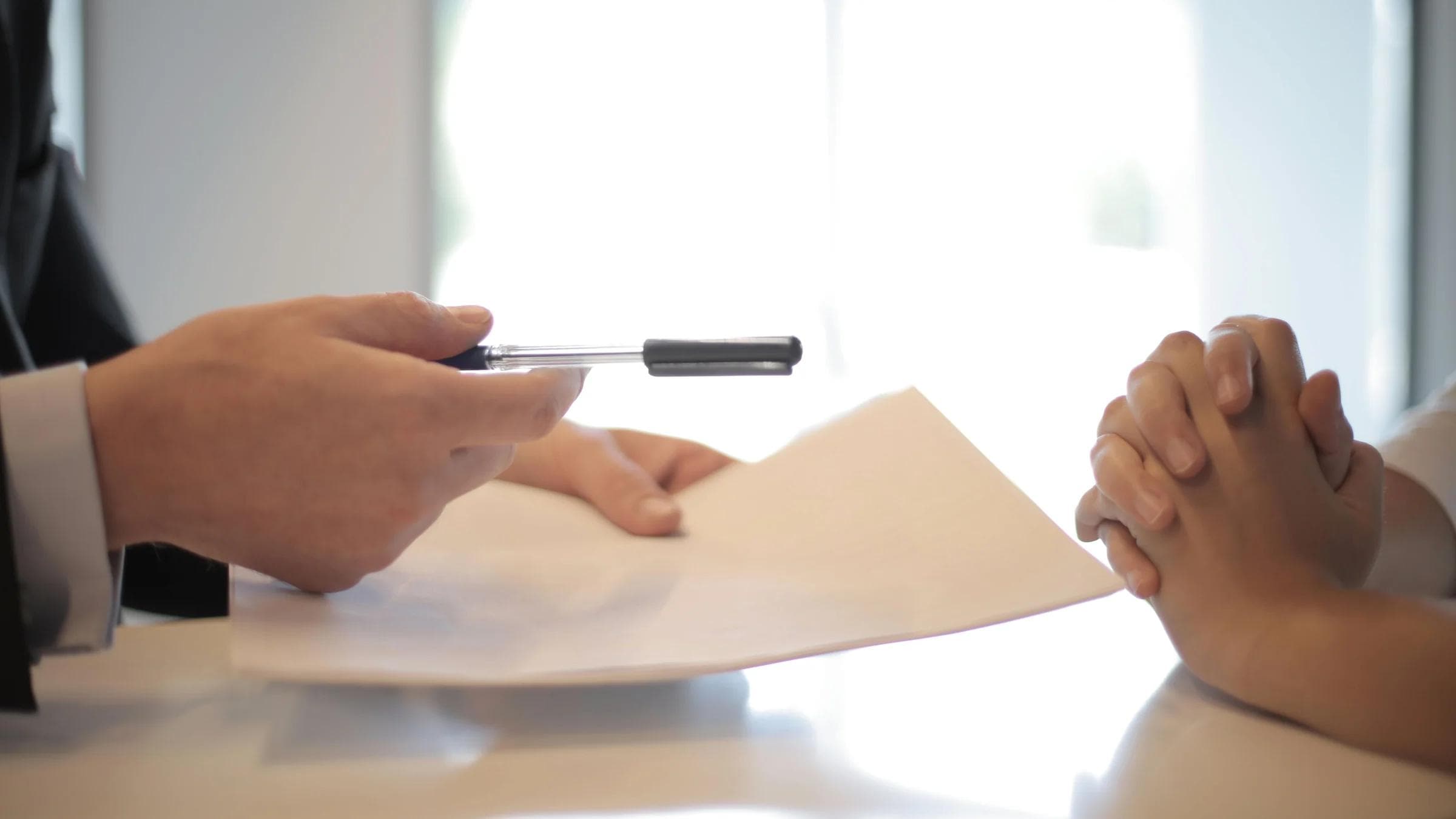 Side view of faceless formal man giving pen and paper to focused female with clenched hands at table on meeting.