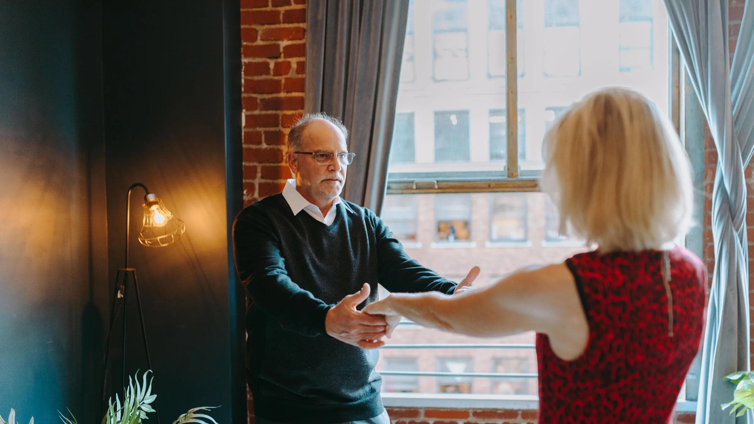 An elderly couple enjoys a dance together in a warmly lit urban apartment, capturing a moment of affection and joy.
