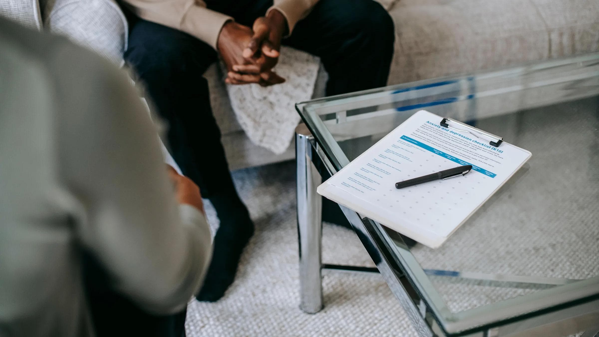From above of unrecognizable ethnic male person in clasped hands sitting near anonymous psychologist in office with glass table and clipboard with notes.