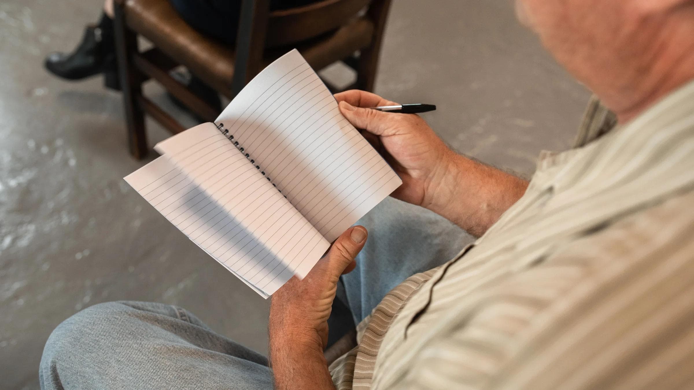 A man writing in a notebook during an indoor meeting or discussion.
