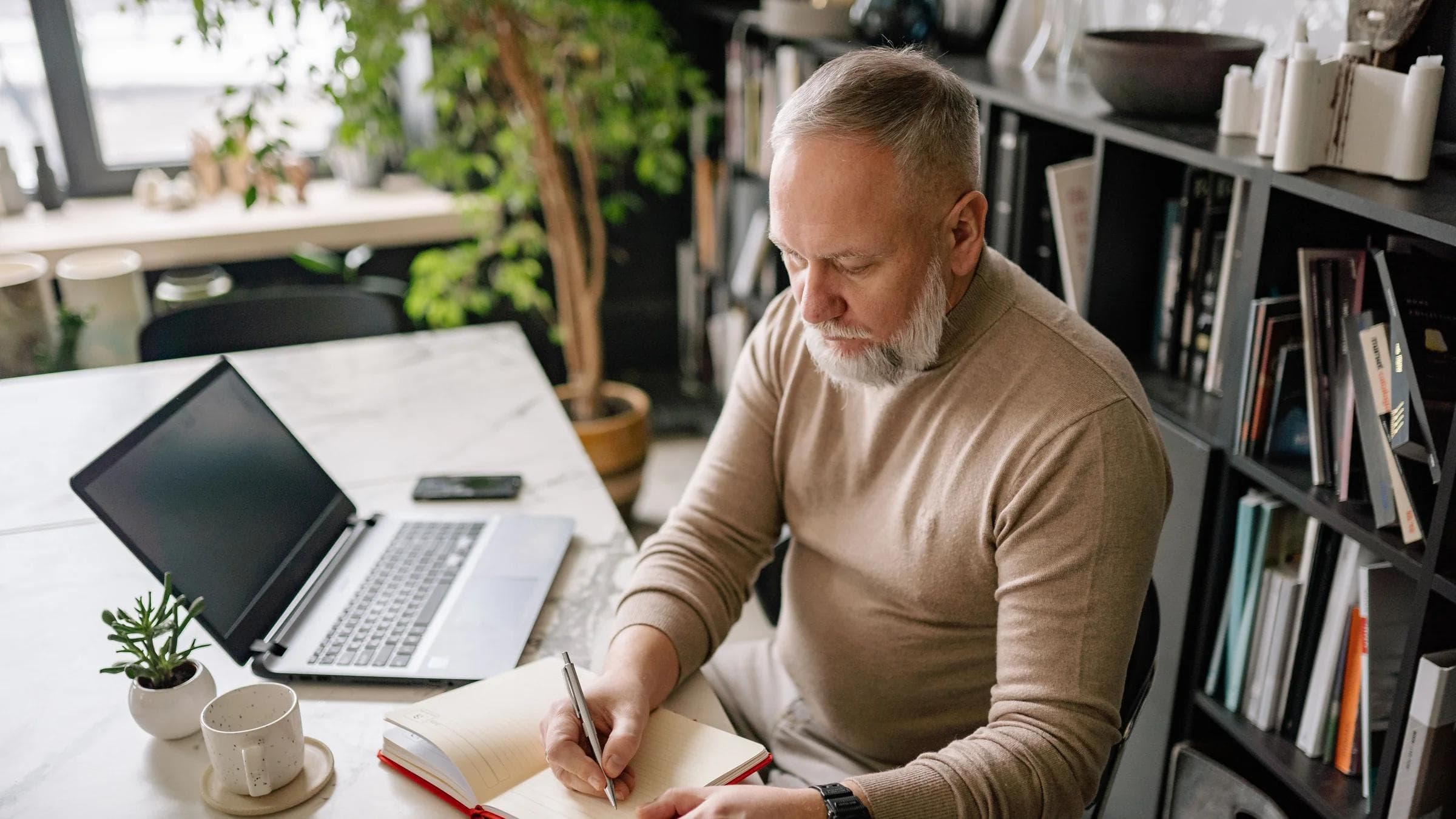 A mature man with a beard writes in a diary at his home office desk next to a laptop.
