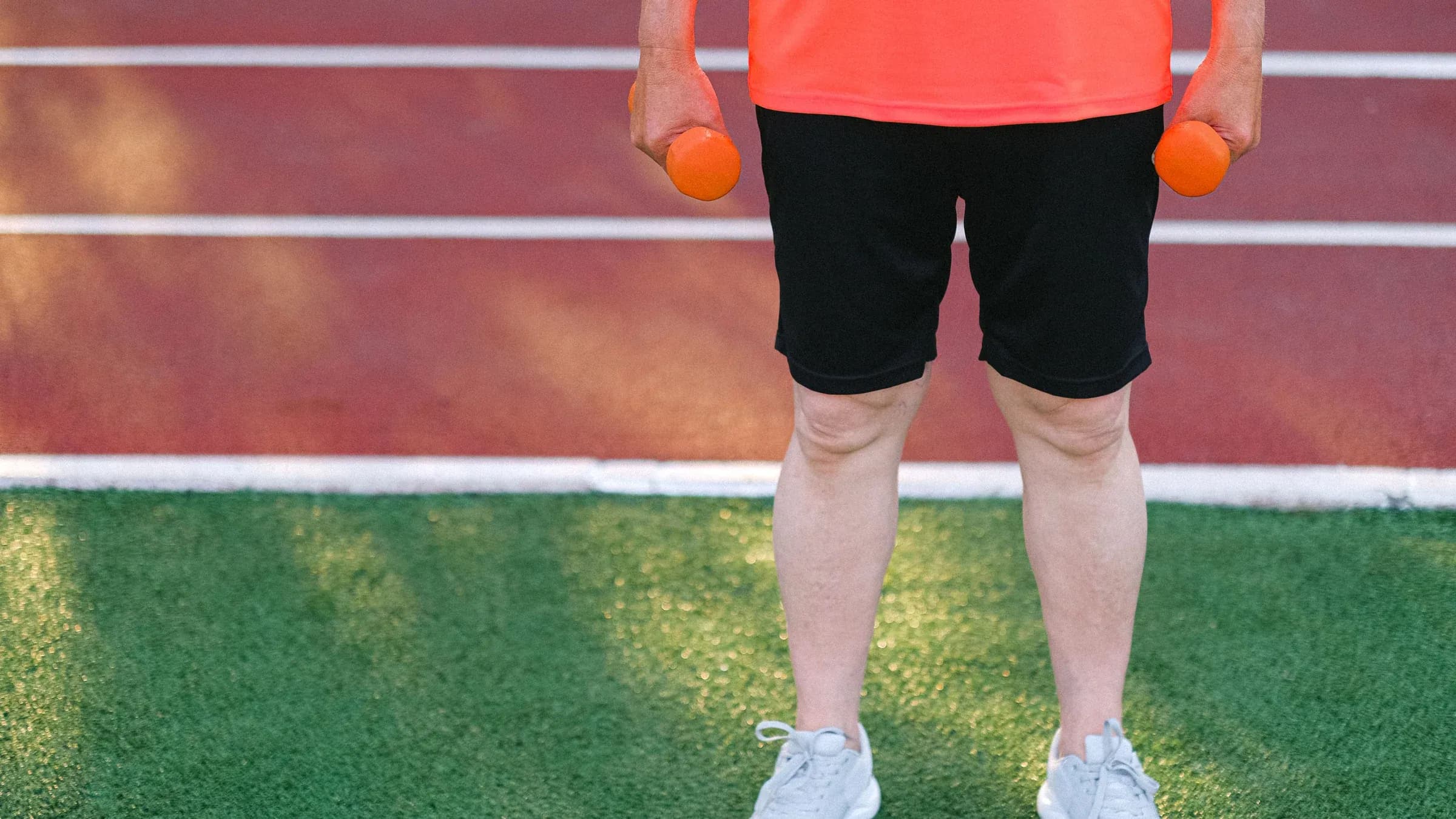 Crop anonymous male showing dumbbells while standing on green grass near racetrack of stadium.